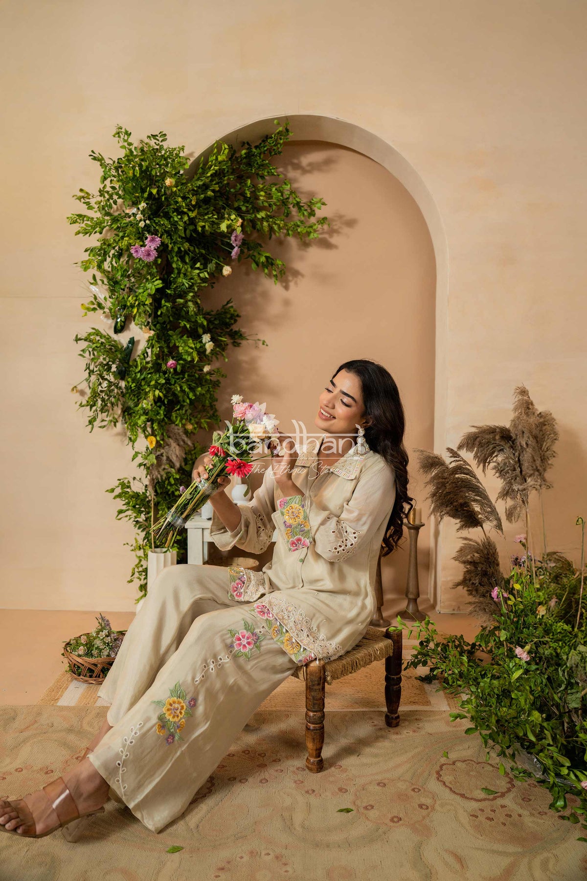 Woman in a floral embroidered outfit sitting in a decorated room with plants and flowers.