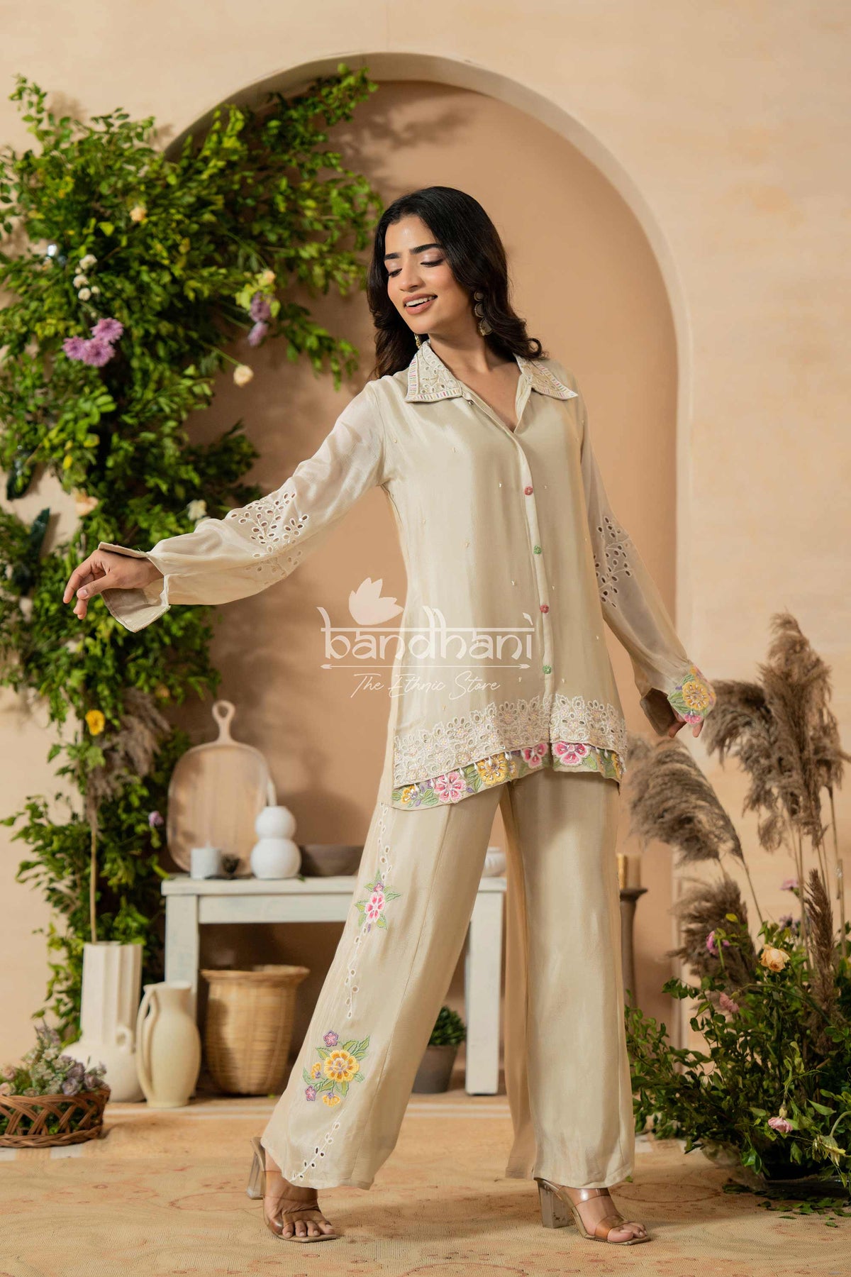 Woman in a beige embroidered outfit standing in a decorative indoor setting with plants and archway.