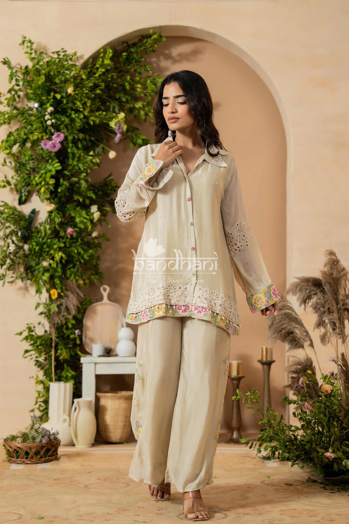 Woman in a beige traditional outfit with floral embroidery in a decorated indoor setting.