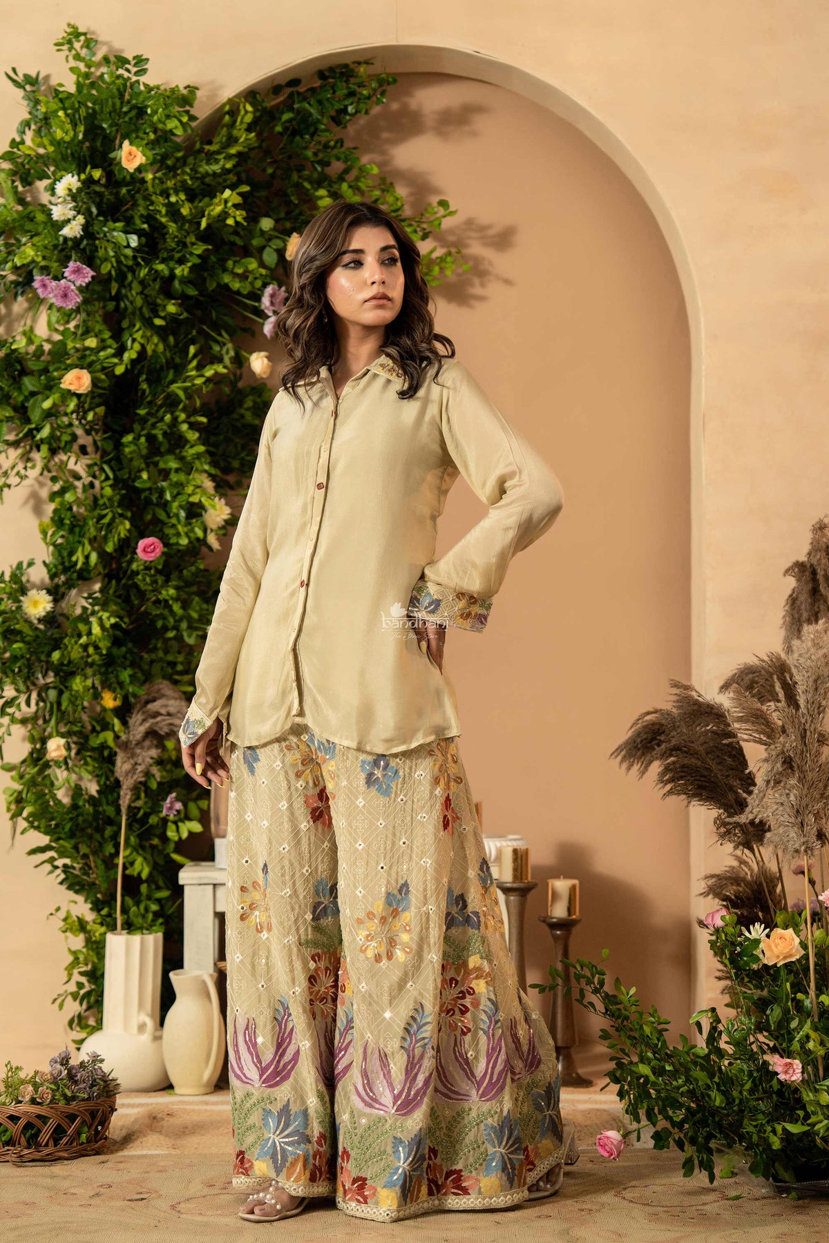 Woman in a light-colored traditional outfit with floral patterns standing in a decorated indoor setting.