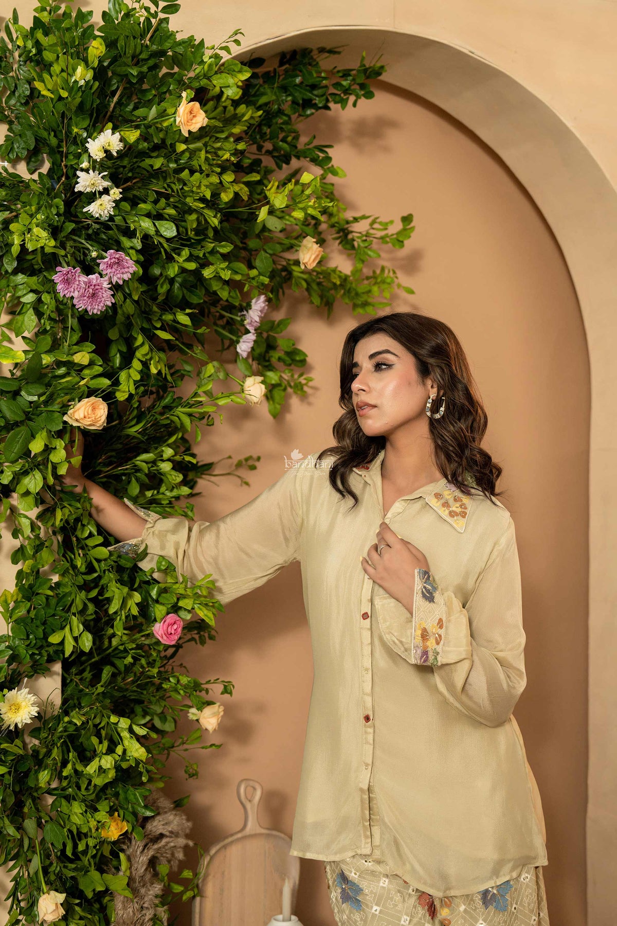 Woman in a light-colored outfit standing next to a floral arrangement against a beige wall.