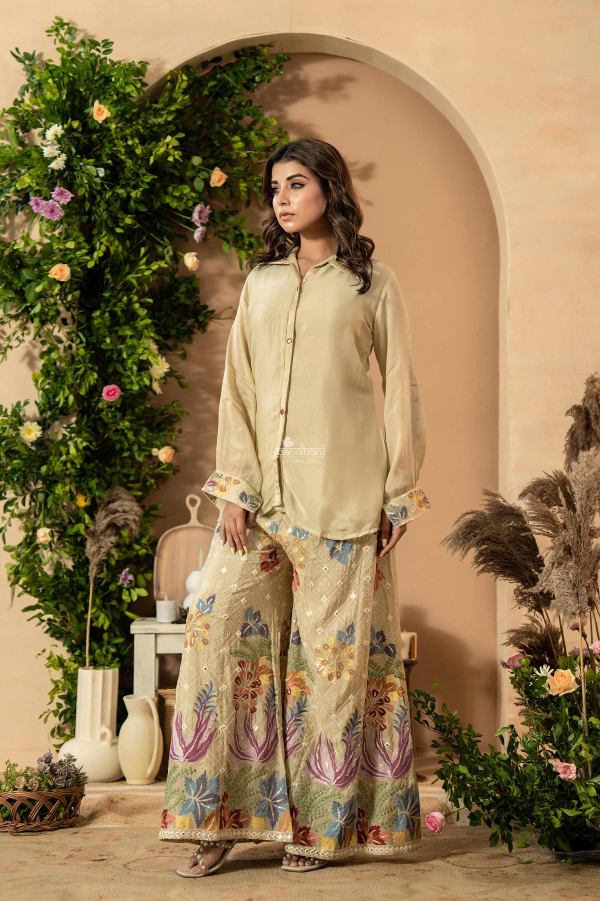 Woman in a light green traditional outfit with floral patterns standing in front of an archway decorated with flowers and plants.