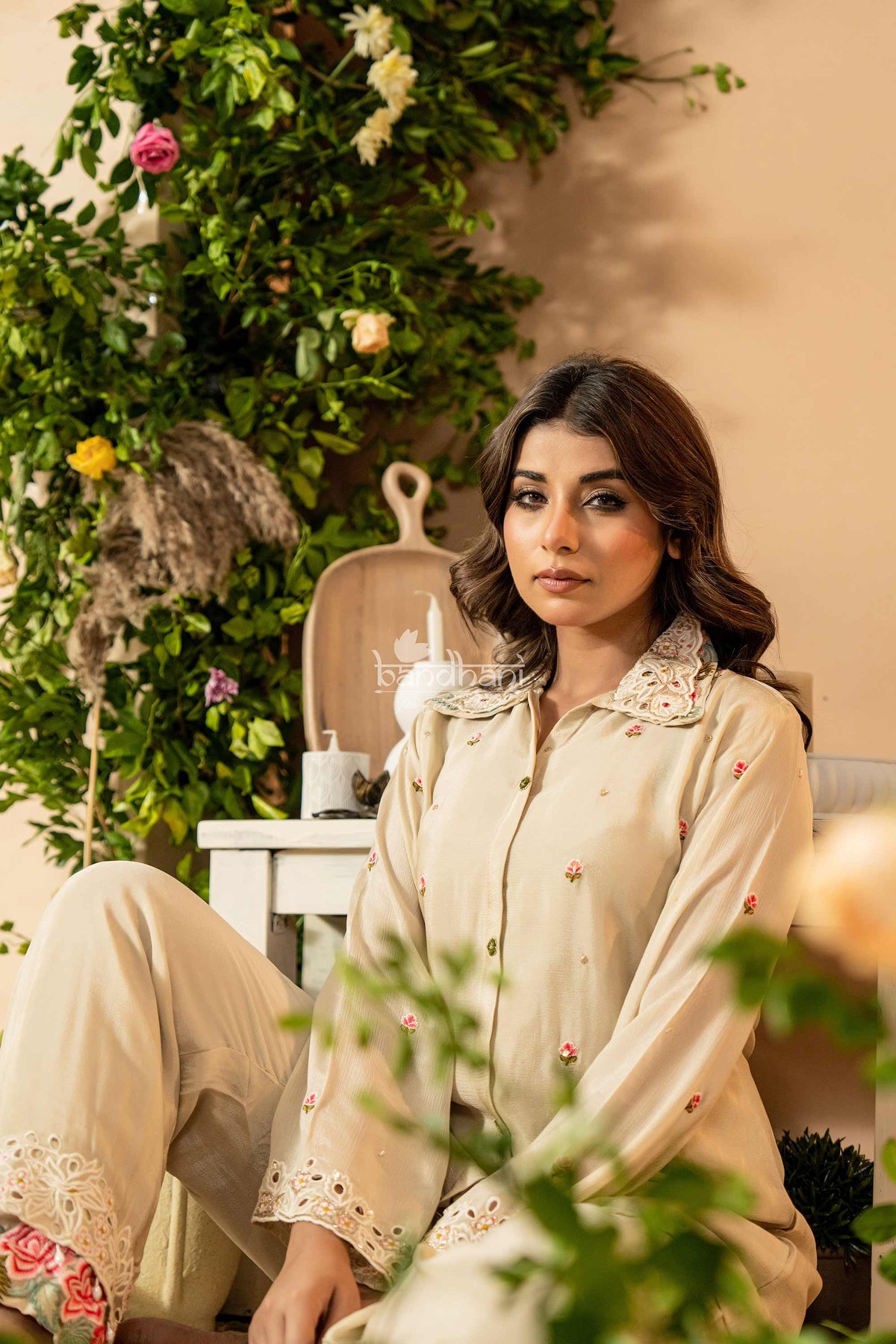 Woman in a beige outfit with floral details sitting in a decorated indoor setting.
