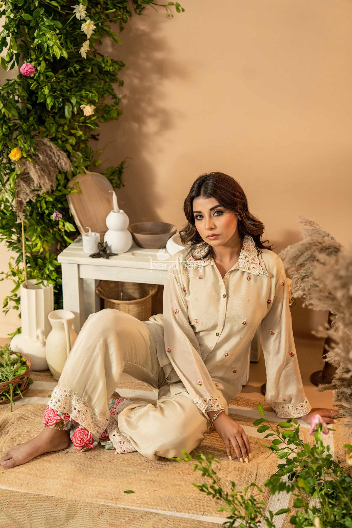 Woman in a cream-colored outfit sitting on a mat with decorative elements and plants.