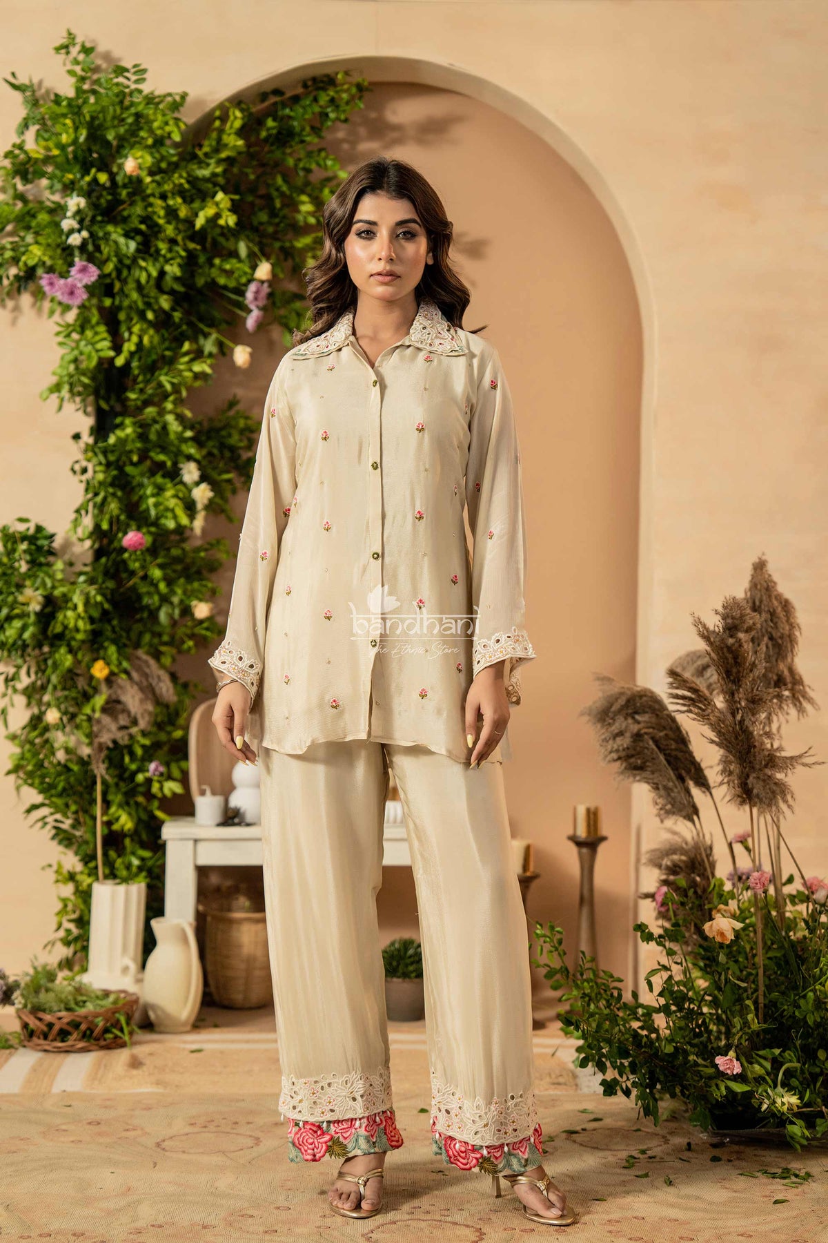 Woman wearing a cream-colored traditional outfit with floral decorations in a decorated indoor setting.