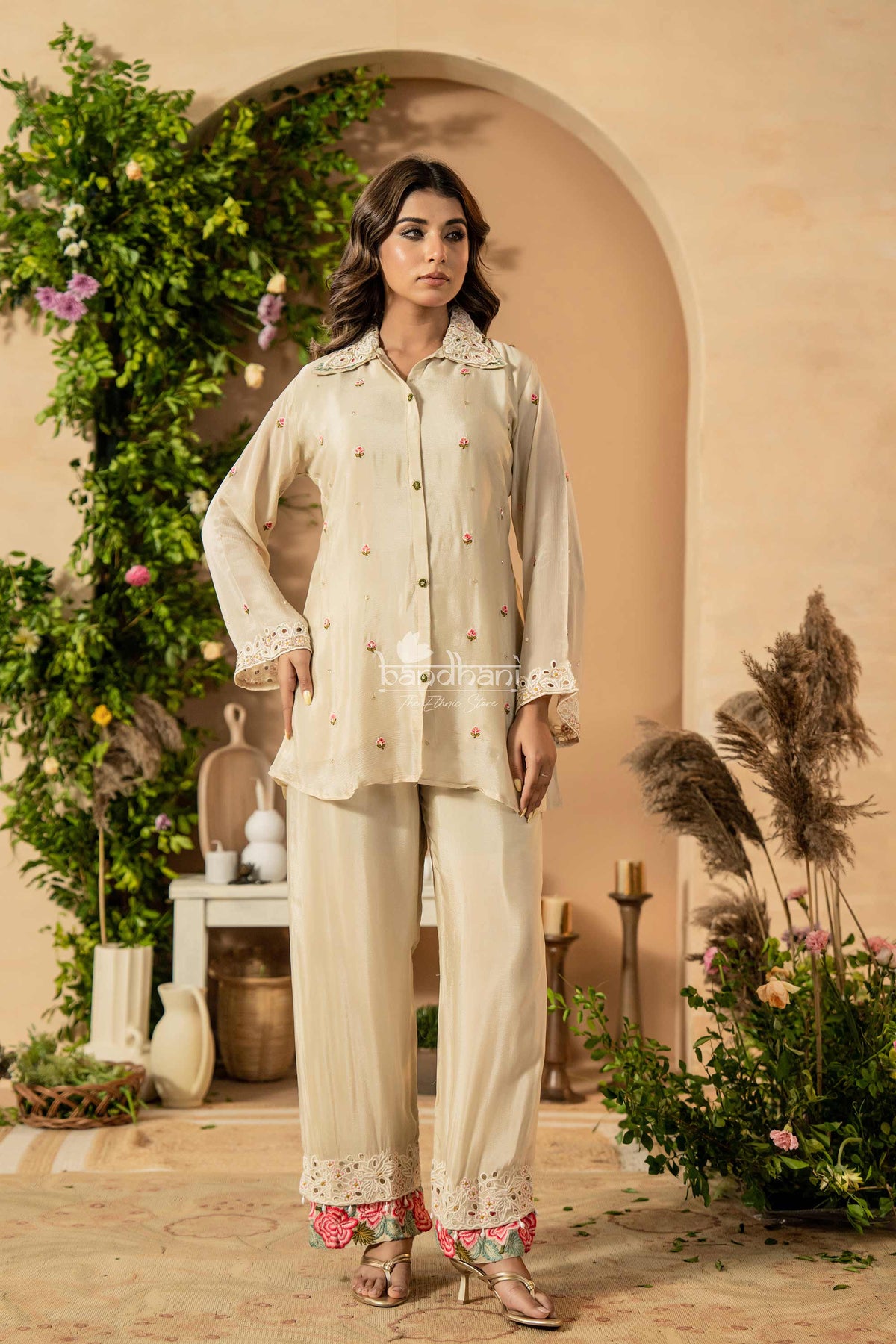 Woman in a traditional outfit standing in a decorated outdoor setting with plants and archway.