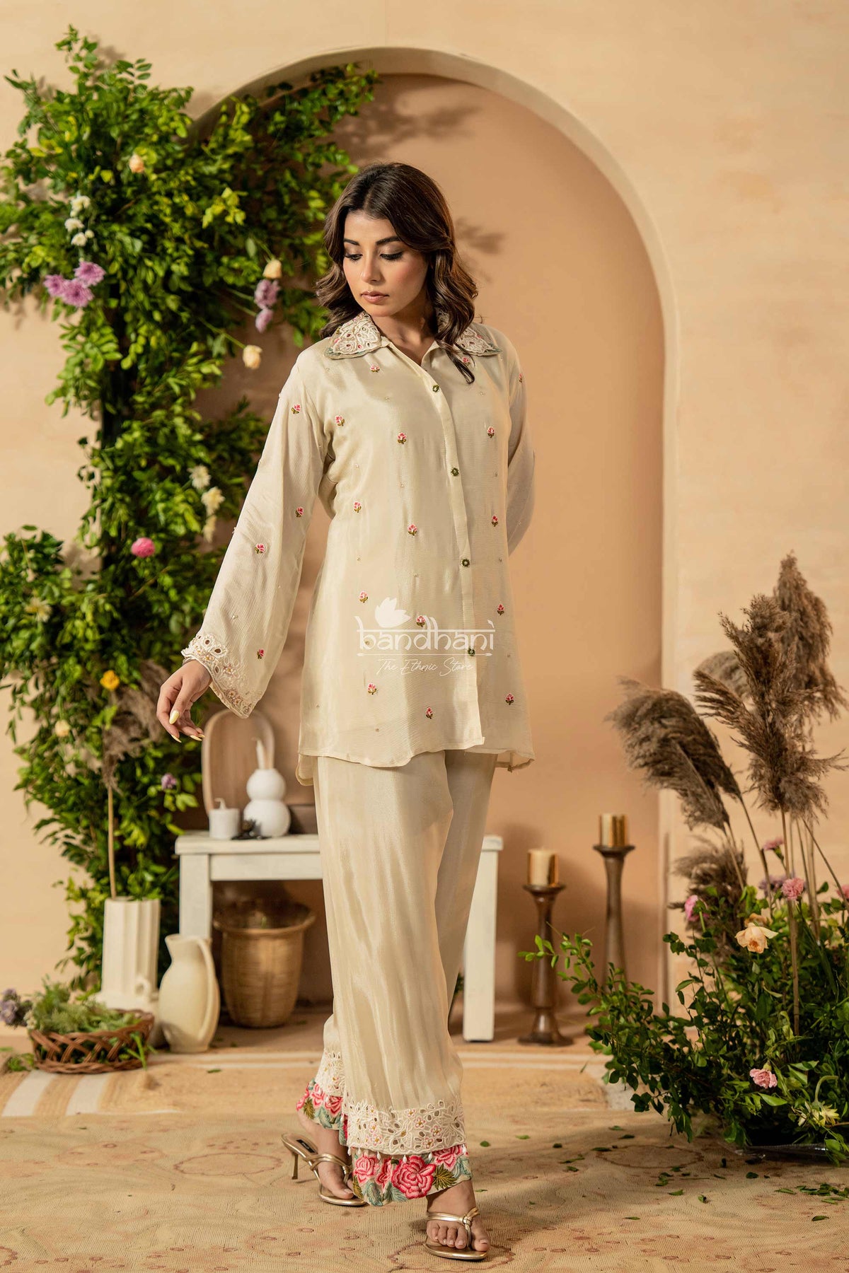Woman in a light-colored traditional outfit standing in a decorative indoor setting with plants and flowers.