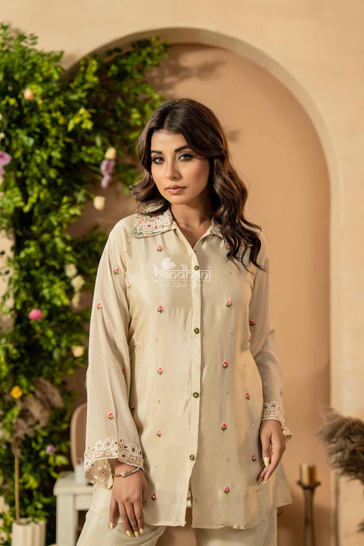Woman wearing a beige traditional outfit with floral decorations in an outdoor setting.