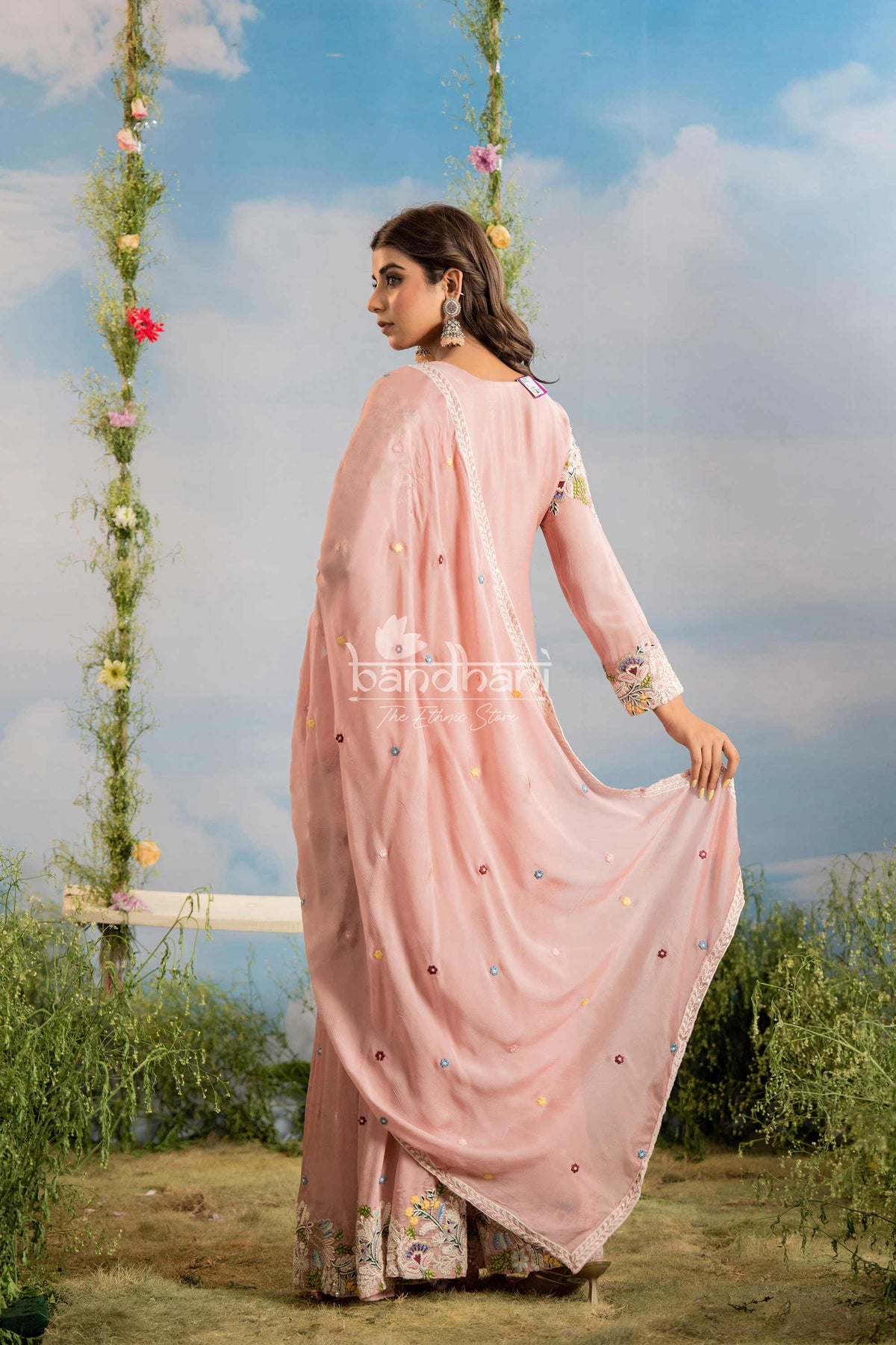 Woman in a pink saree standing on a swing with floral decorations against a blue sky.