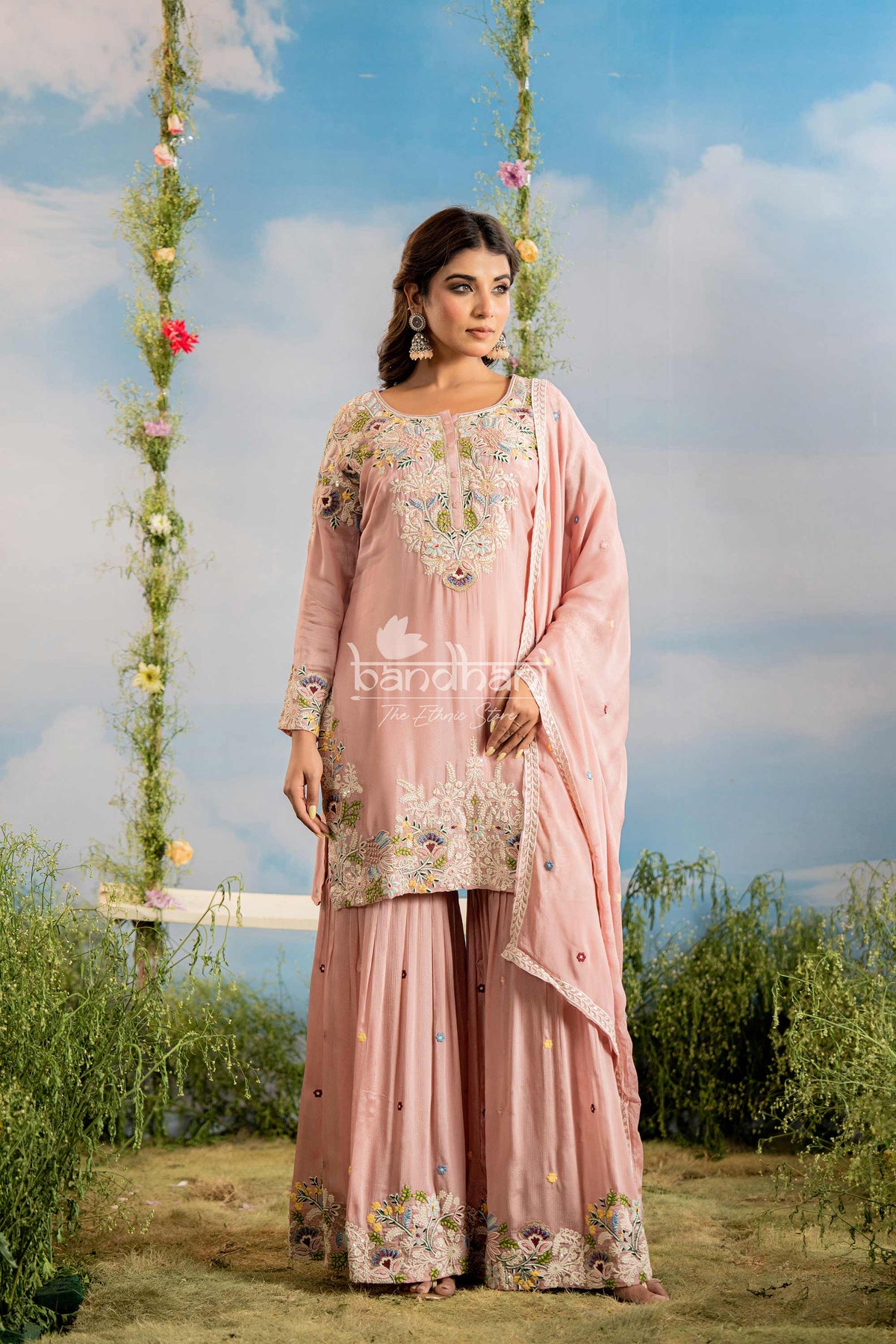Woman in a pink traditional outfit with floral patterns standing outdoors against a blue sky.
