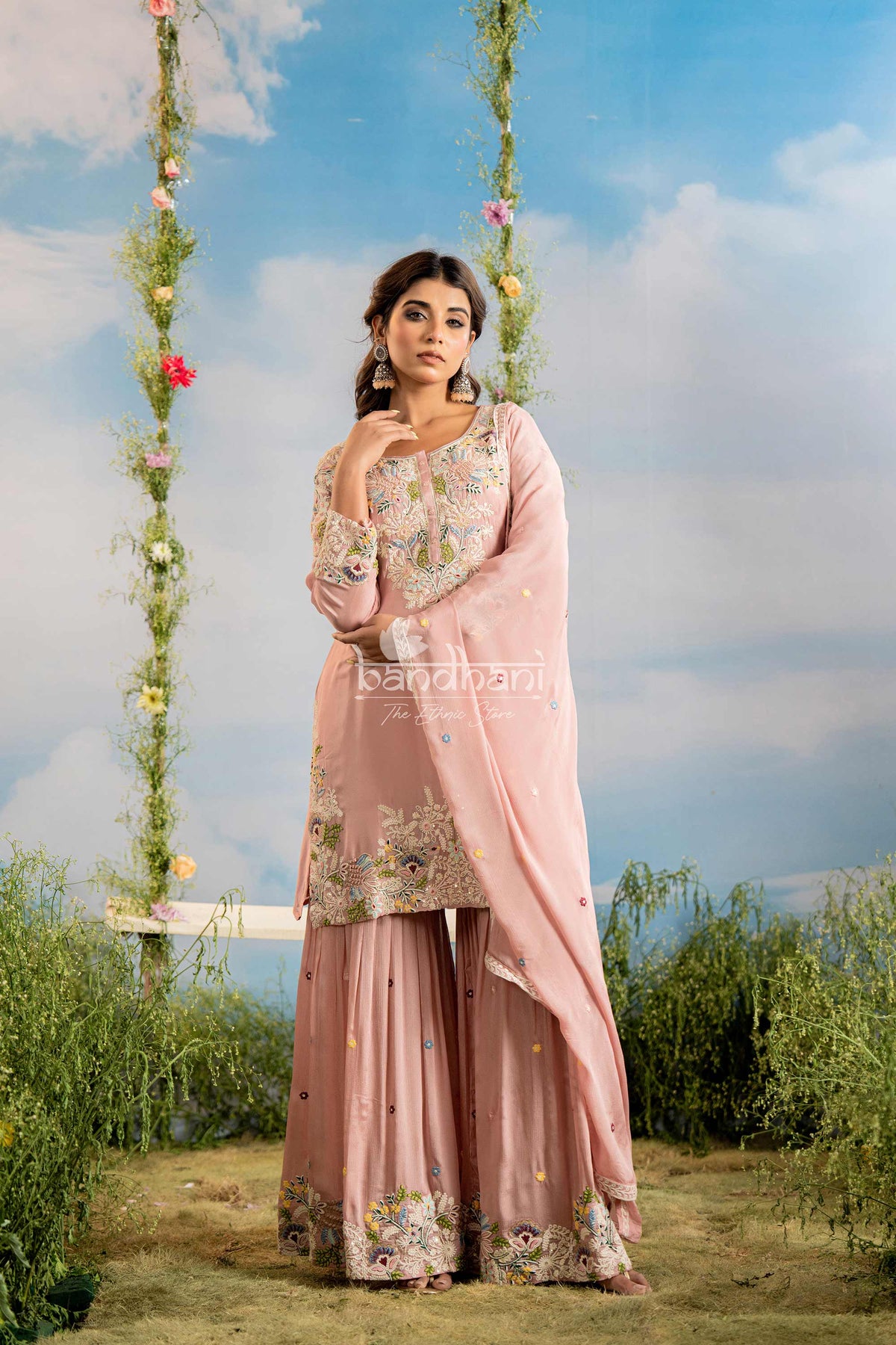 Woman in a pink traditional outfit standing outdoors with greenery and a blue sky.
