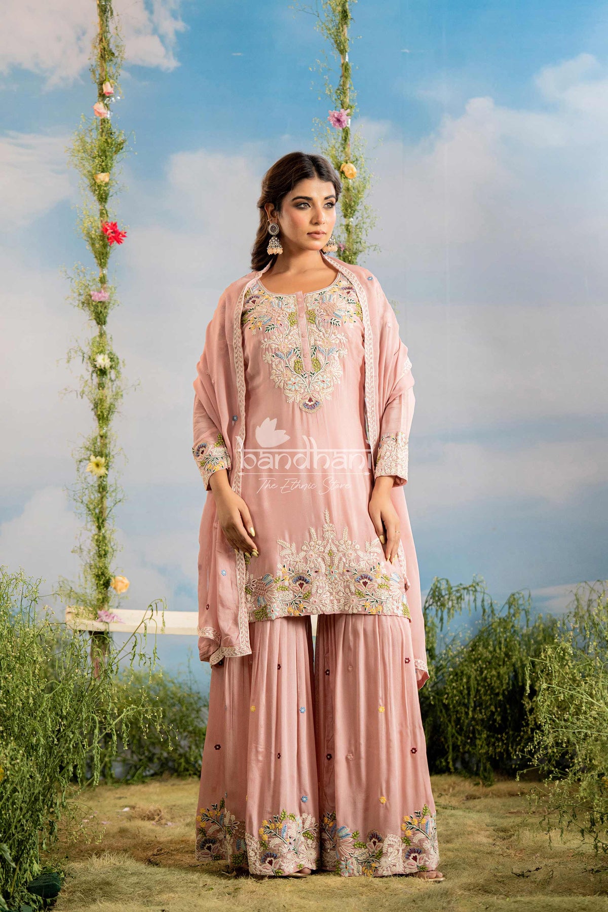 Woman in a pink embroidered outfit standing on a swing with floral decorations against a blue sky.