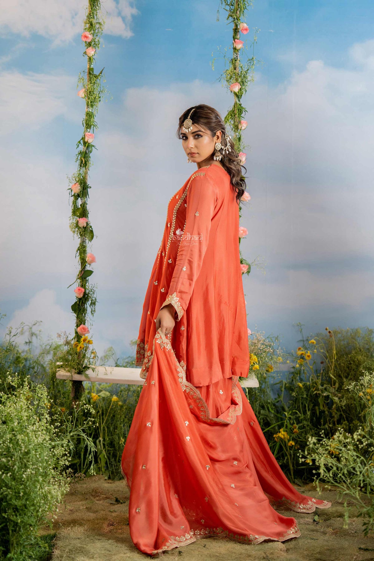 Woman in a coral traditional outfit sitting on a swing with floral decorations against a blue sky.