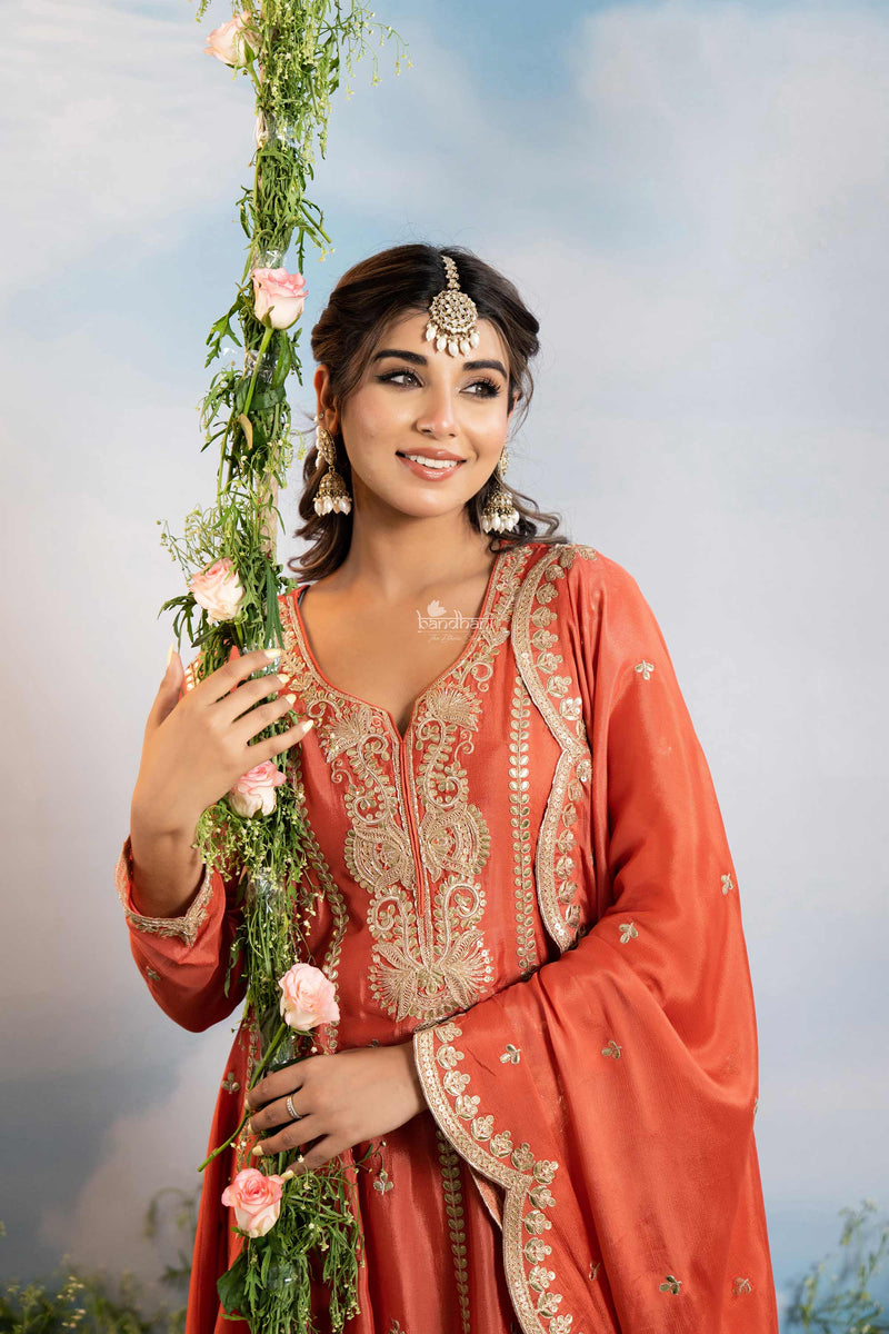 Woman in traditional orange and gold embroidered outfit holding floral decorations against a blurred natural background