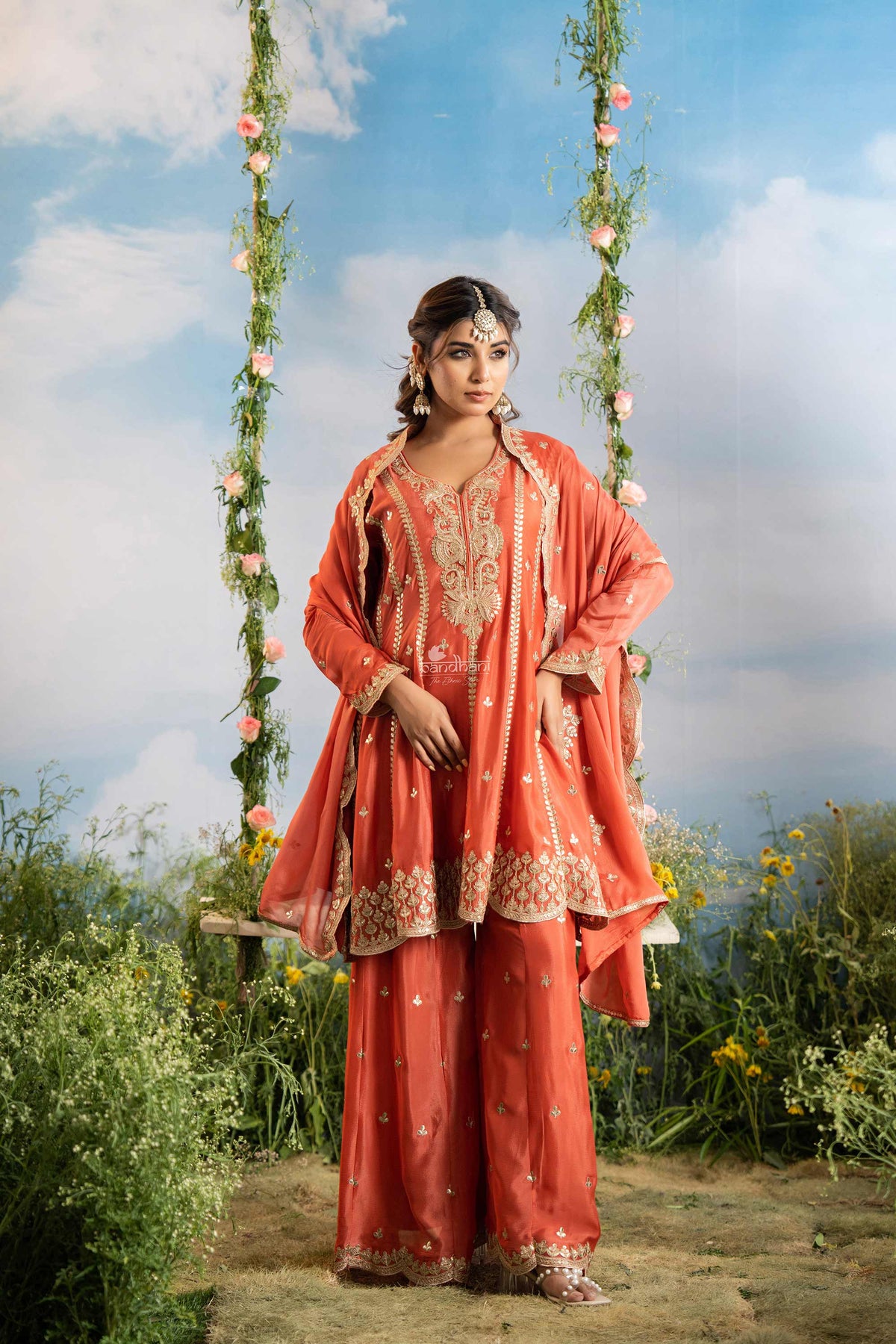 Woman in traditional coral outfit with floral decorations on a swing against a blue sky.