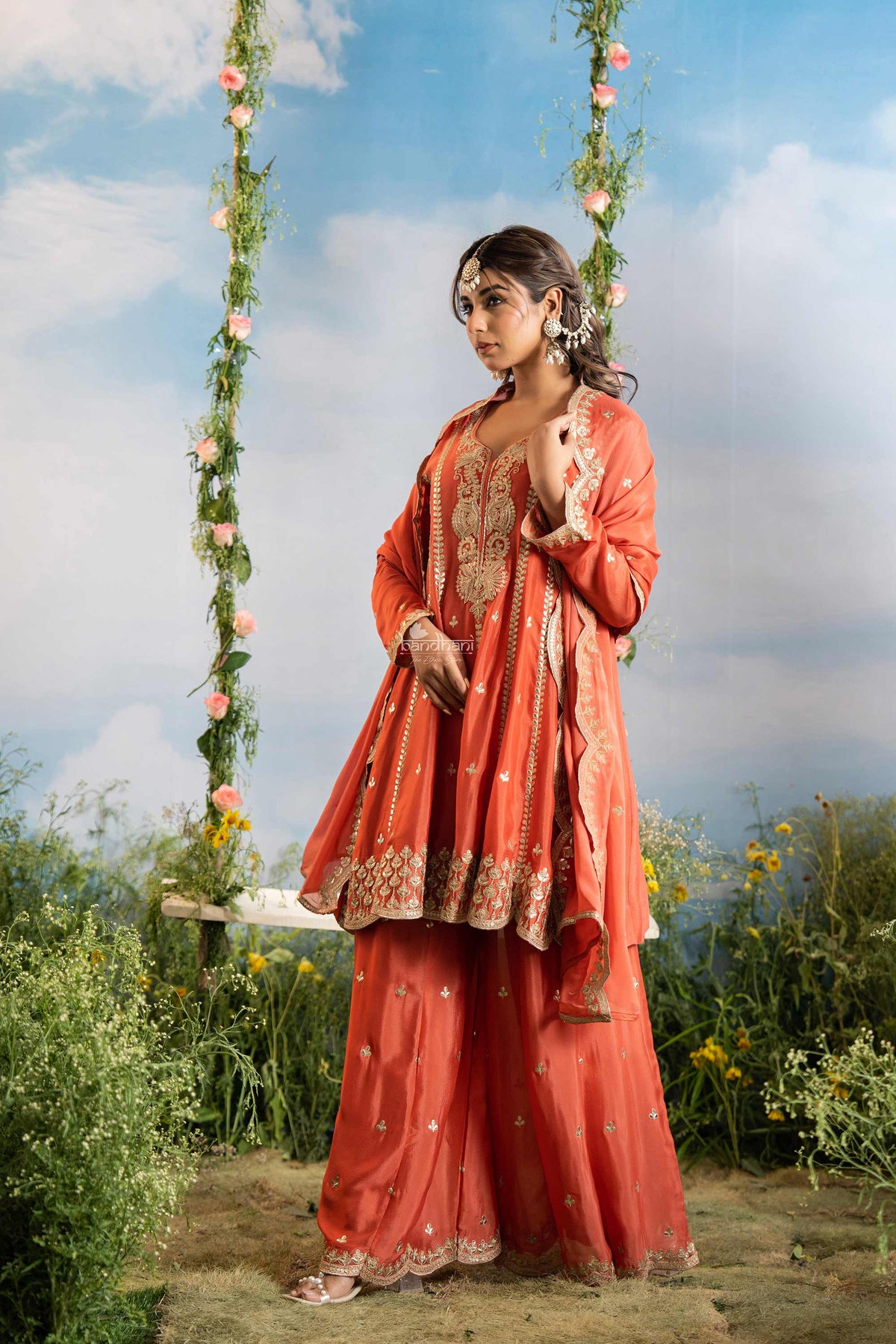 Woman in an orange traditional outfit standing on a swing with floral decorations against a blue sky.