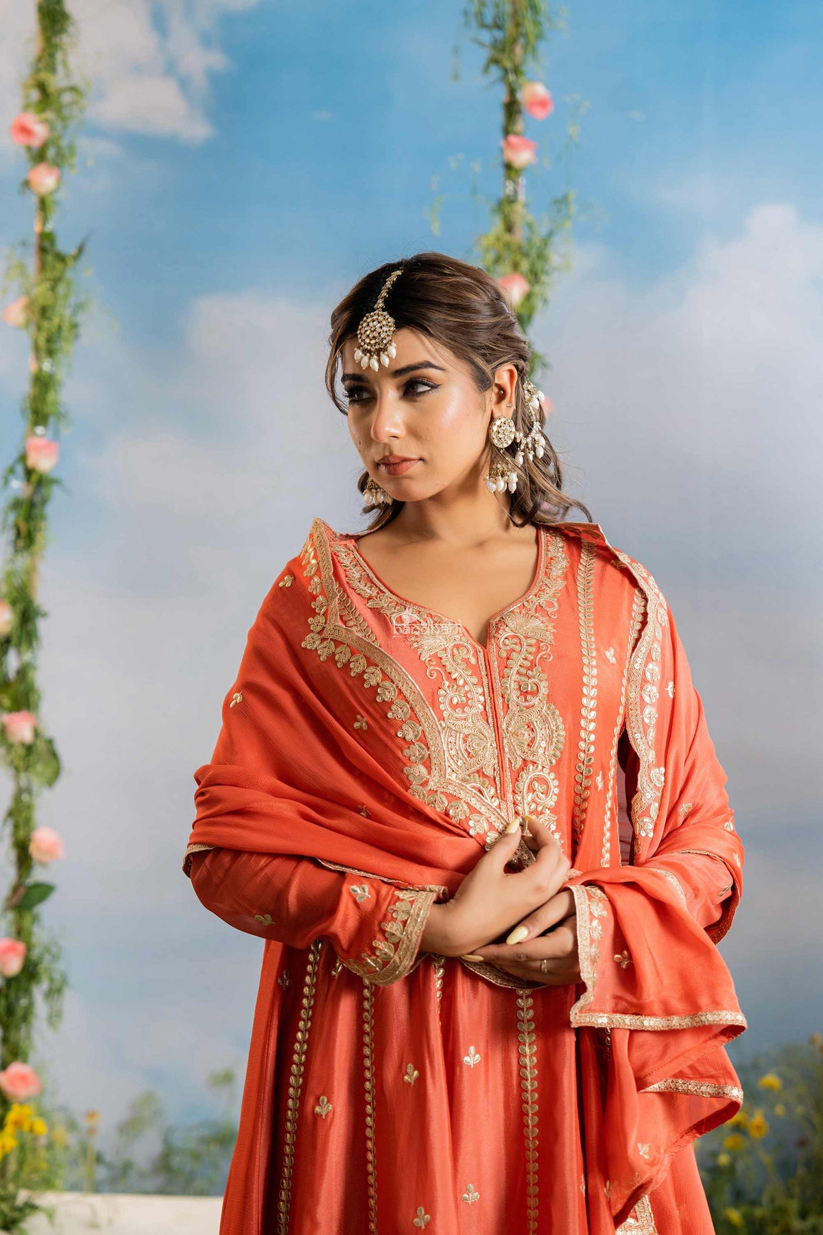 Woman in traditional orange outfit with floral decorations against a blue sky.