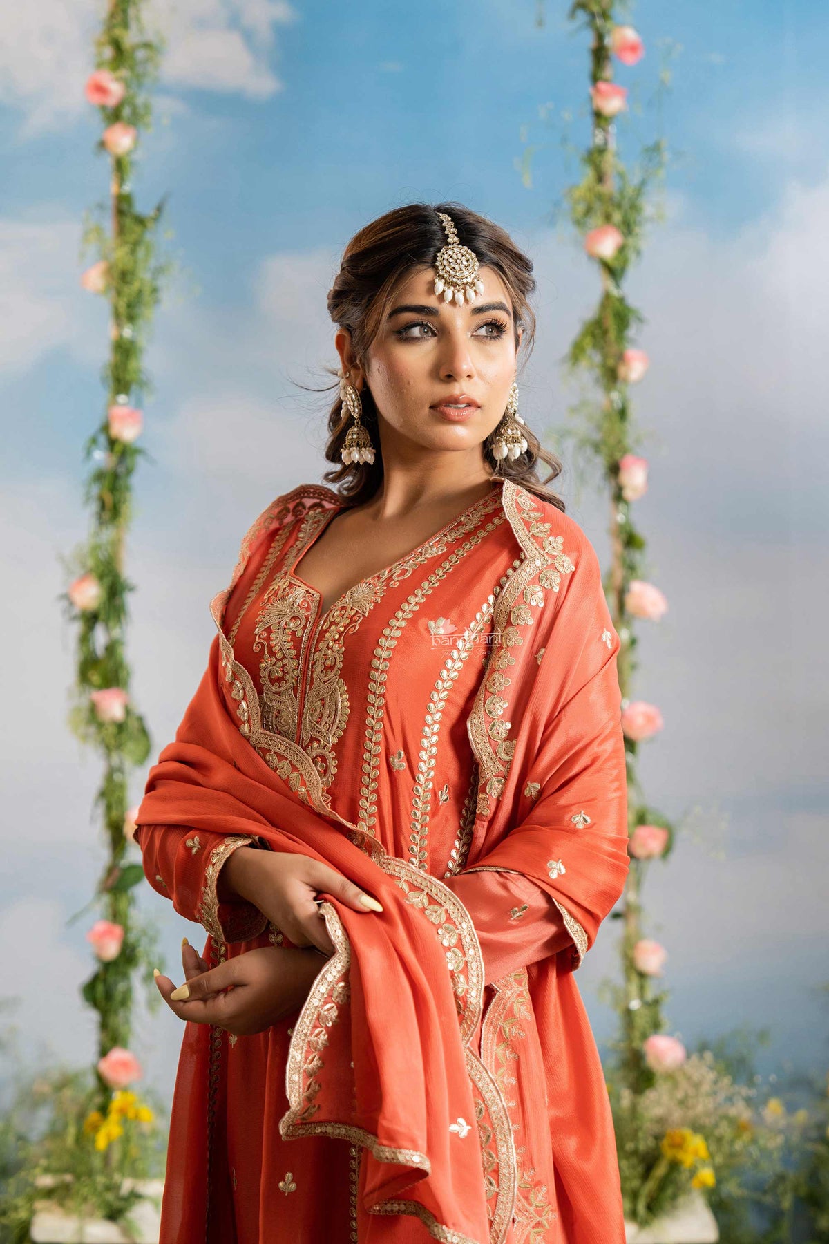 Woman in traditional orange outfit with gold embroidery against a floral swing backdrop