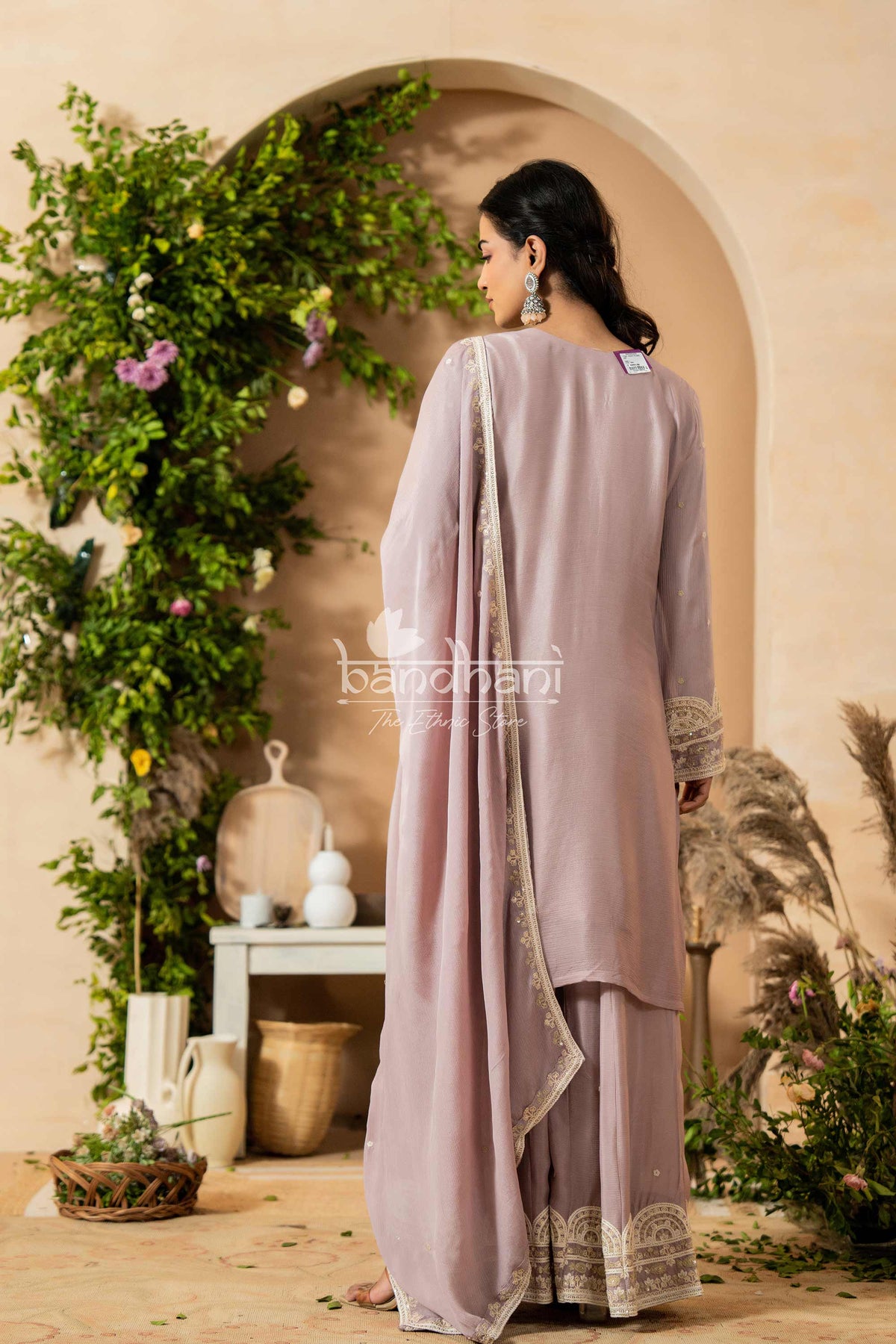 Woman in a light purple traditional outfit standing in a decorated room with plants and decor.