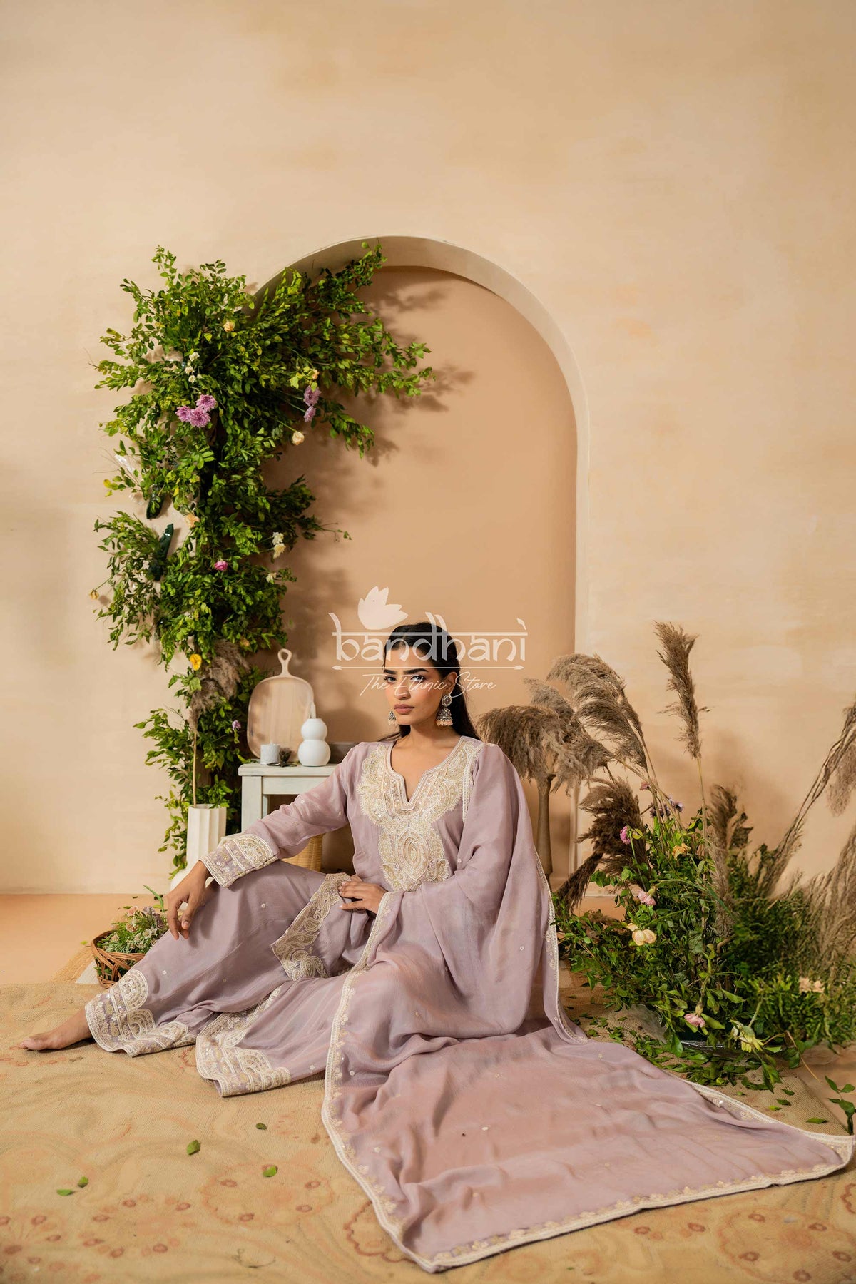 Woman in a lavender traditional outfit sitting in a decorated room with plants and archway.