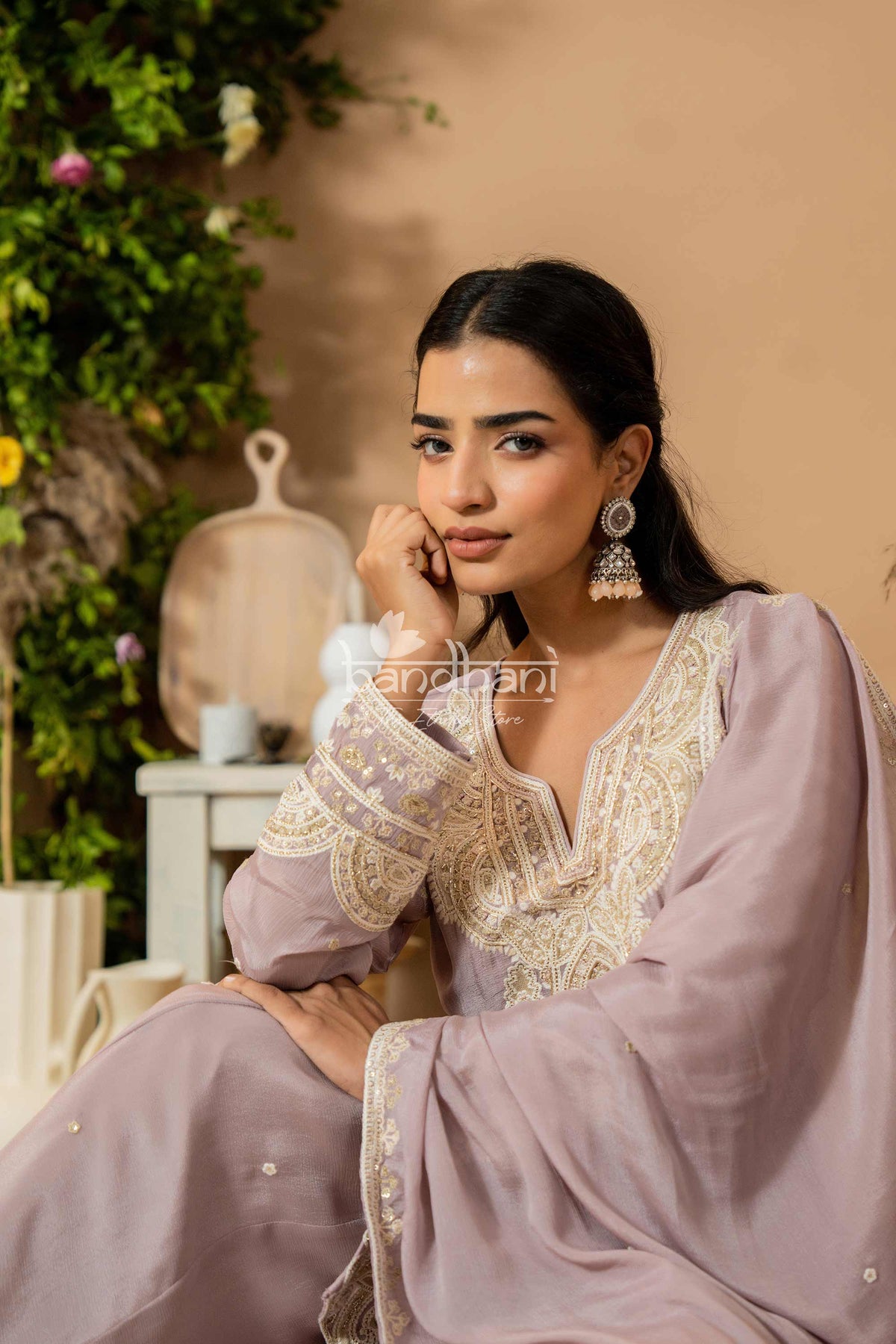 Woman in a lavender traditional outfit with intricate embroidery, sitting in a decorative setting.