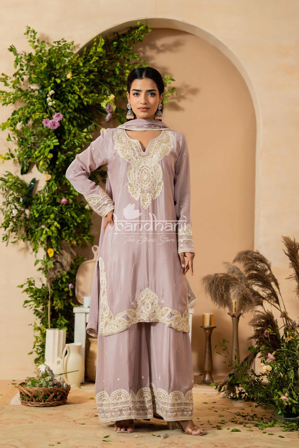 Woman in a lavender traditional outfit with intricate white embroidery standing in a decorated indoor setting.