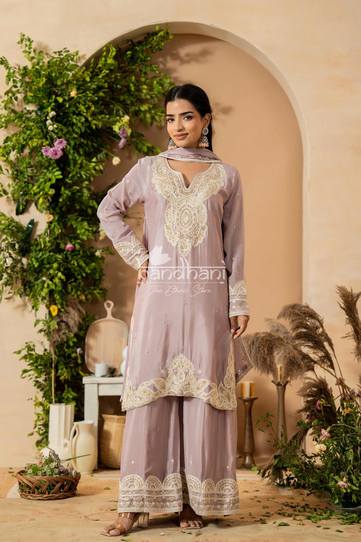 Woman in a lavender traditional outfit with white embroidery standing in a decorative setting with plants and archway.