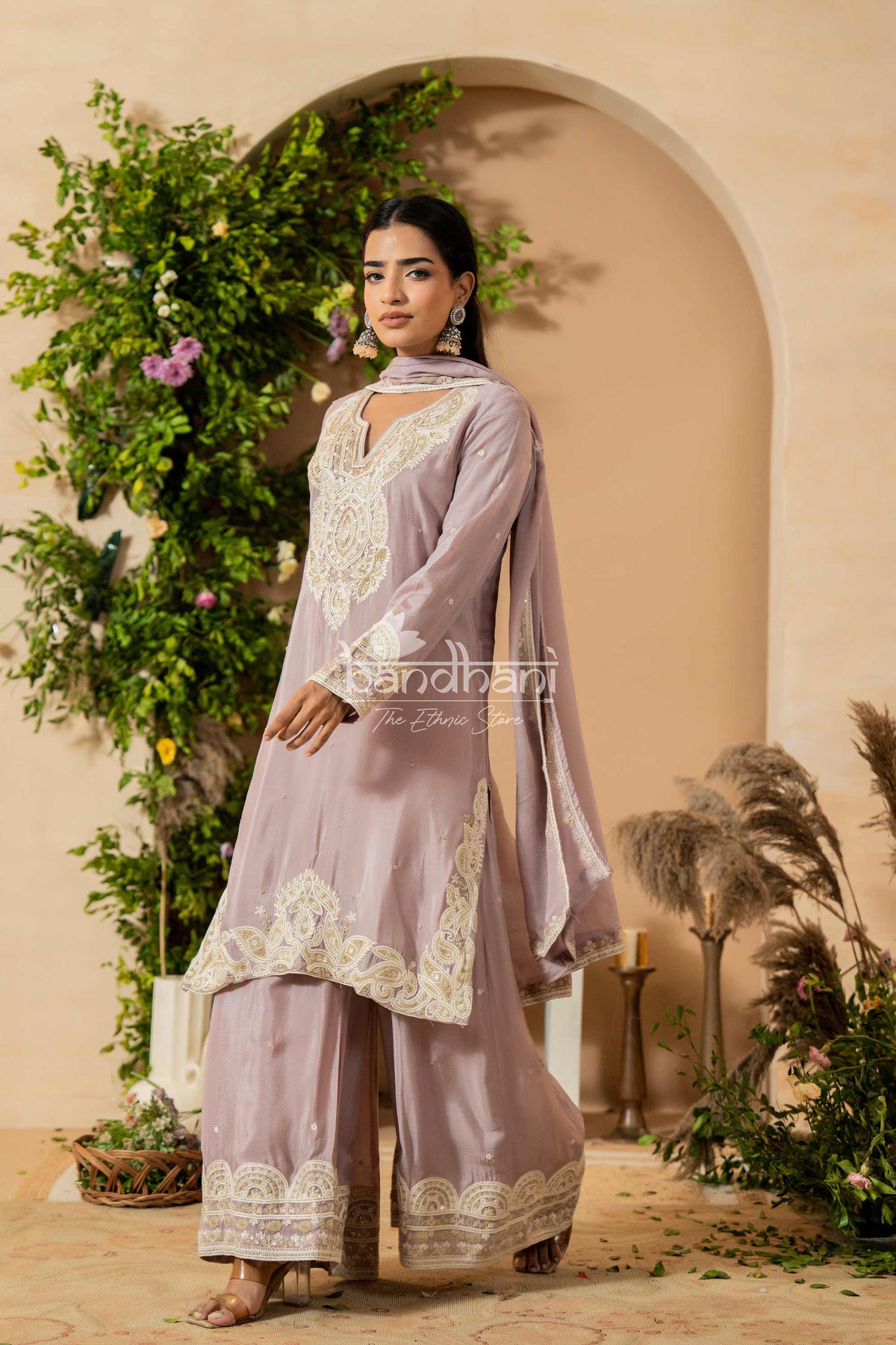 Woman in a lavender traditional outfit with white embroidery standing in a decorative setting.