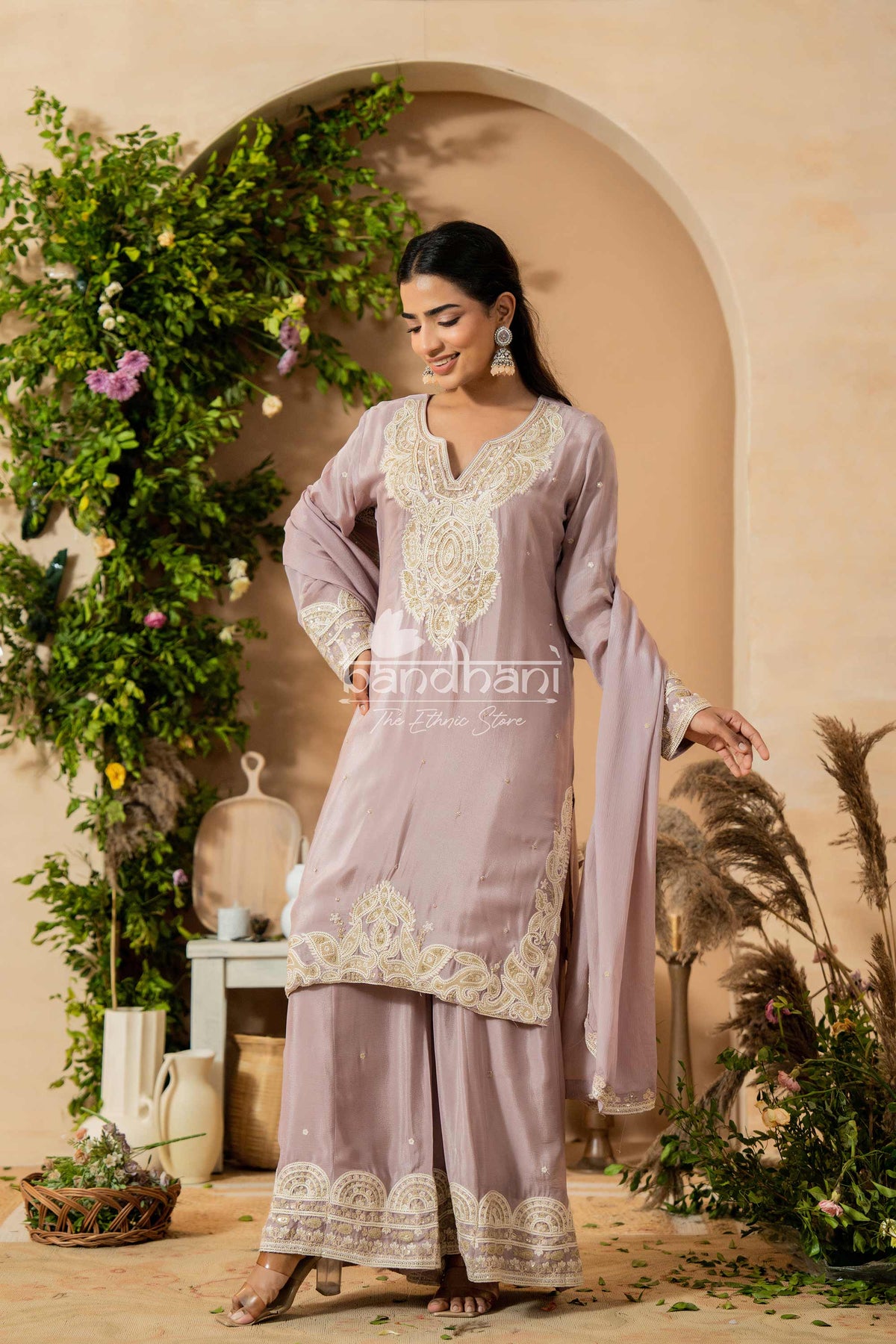 Woman in a lavender traditional outfit with intricate embroidery standing in a decorated outdoor setting.