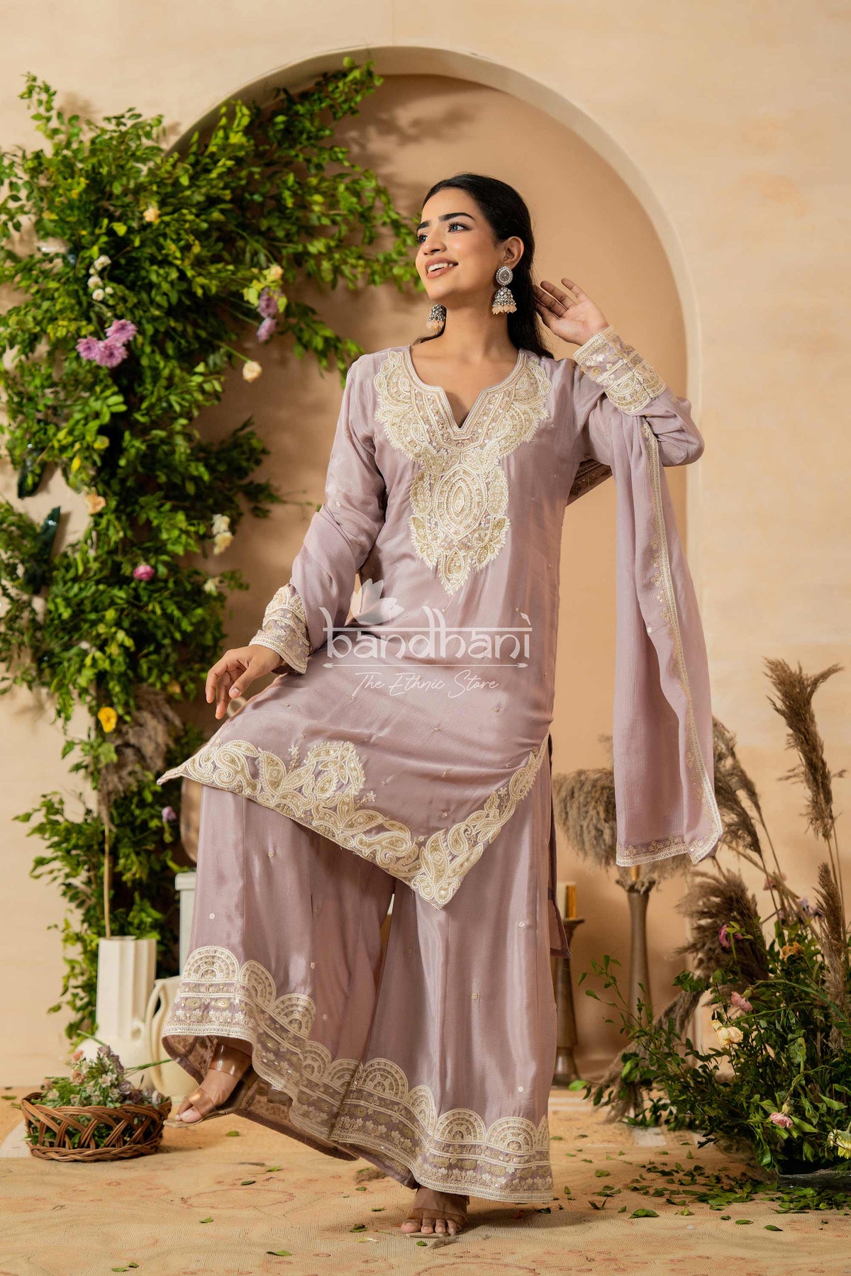 Woman in a lavender traditional outfit with white embroidery standing in a decorated indoor setting.