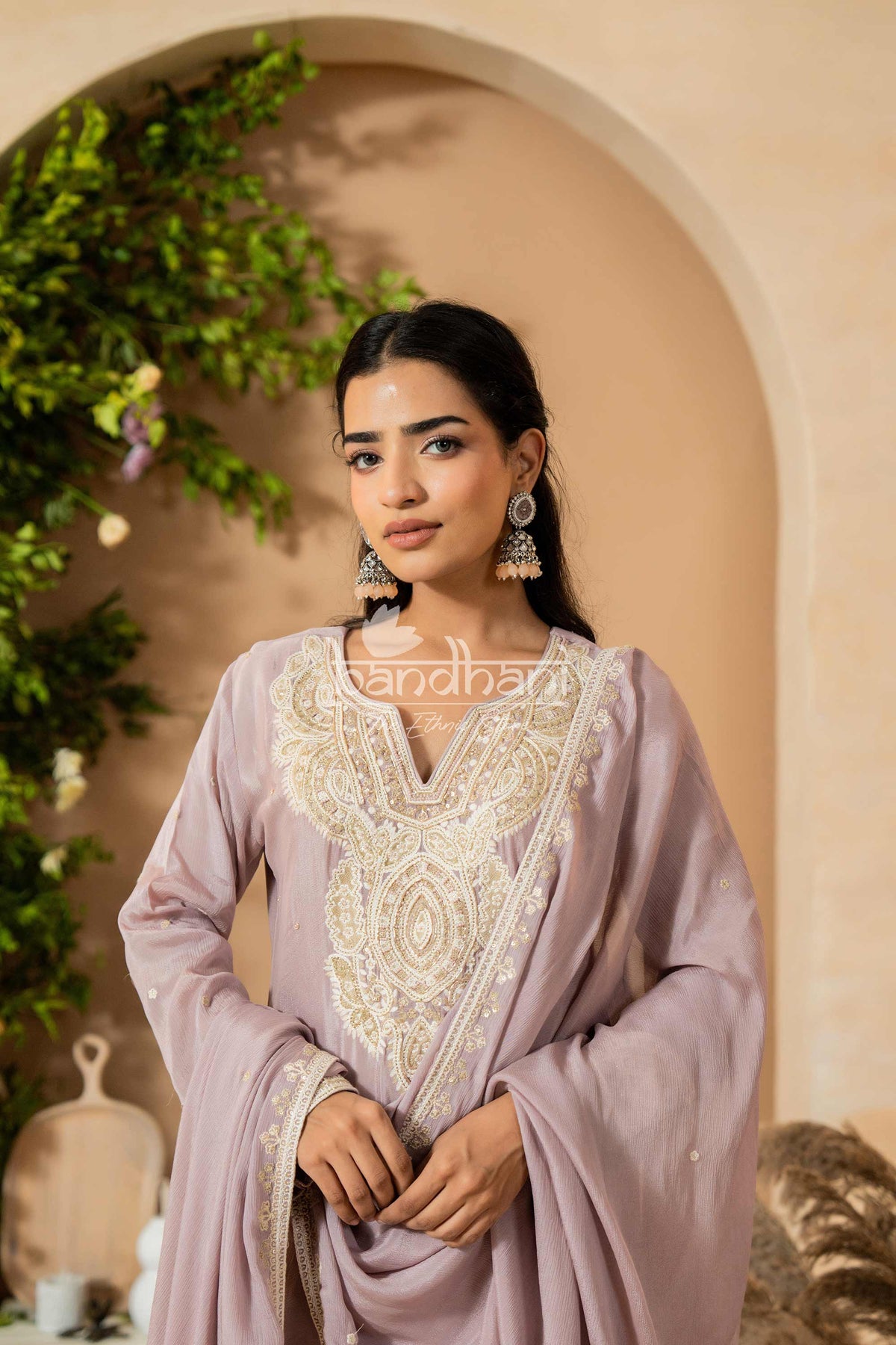 Woman wearing a light purple traditional outfit with intricate white embroidery, standing against a beige wall with greenery.