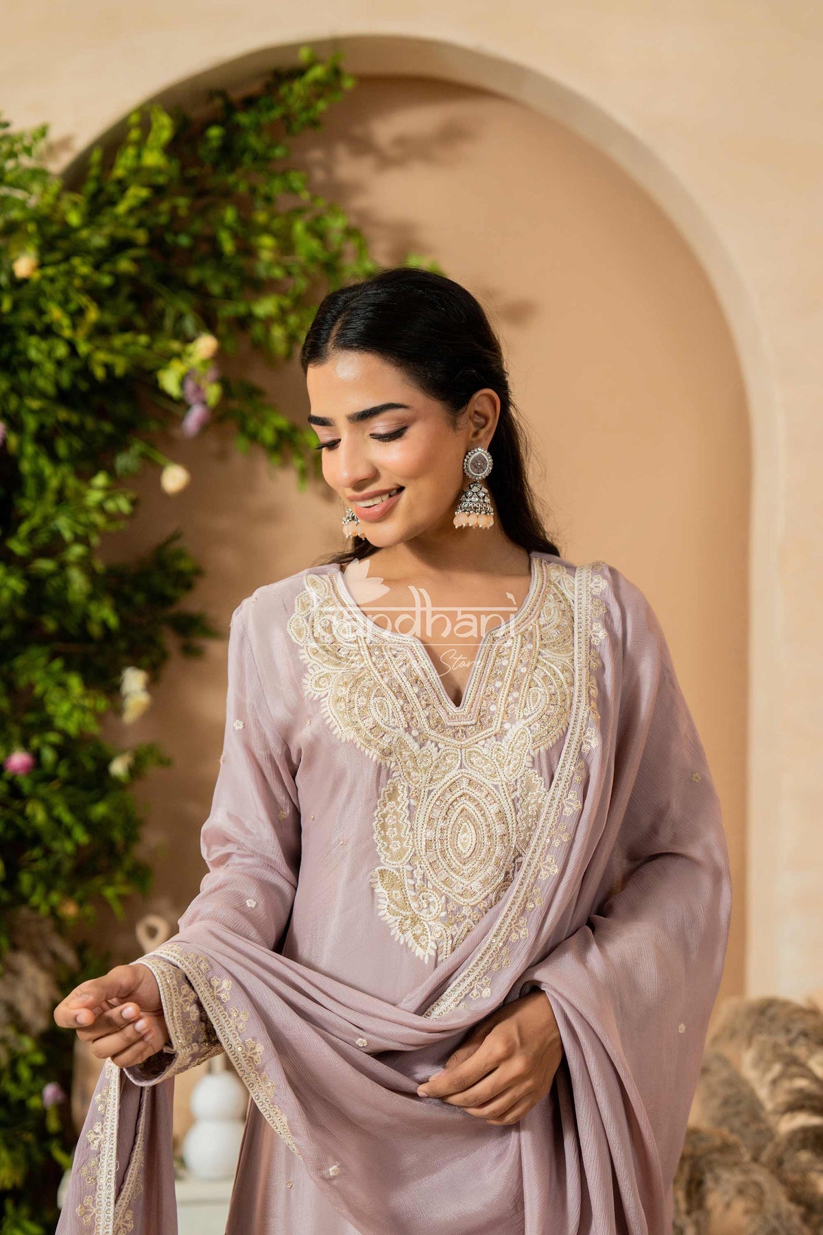Woman wearing a light purple traditional outfit with intricate embroidery, standing against a decorative archway.