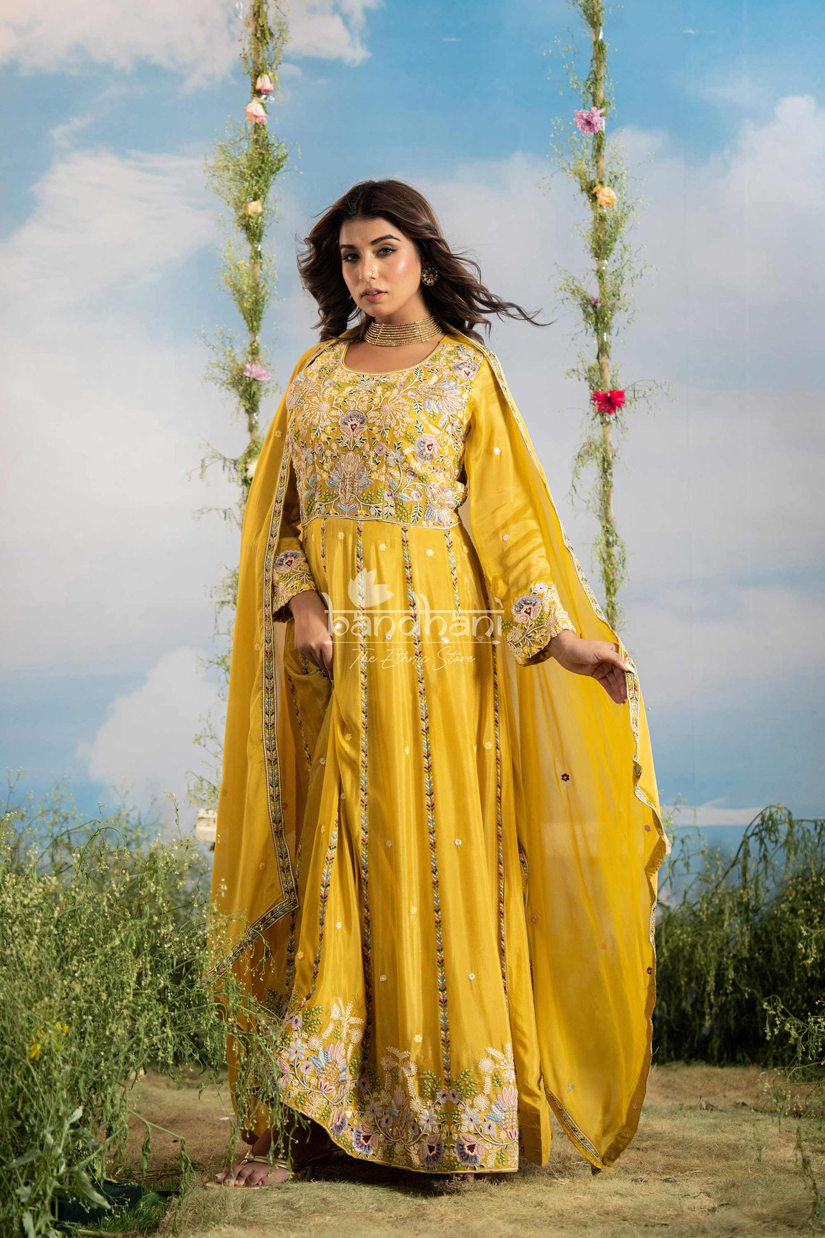 Woman in a yellow traditional outfit with floral patterns, standing outdoors with a scenic background.