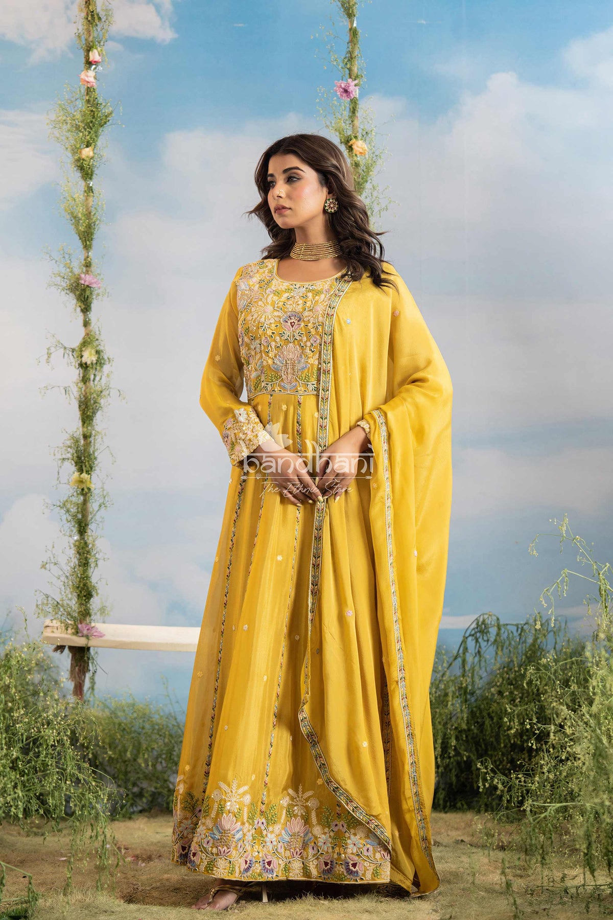 Woman in a yellow traditional outfit with floral patterns, standing outdoors with greenery and a blue sky.