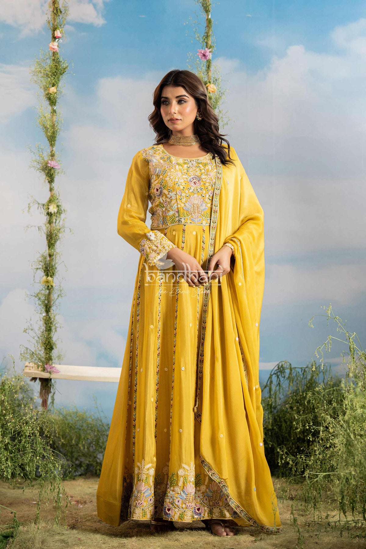 Woman in a yellow traditional outfit with floral patterns sitting on a swing against a blue sky.