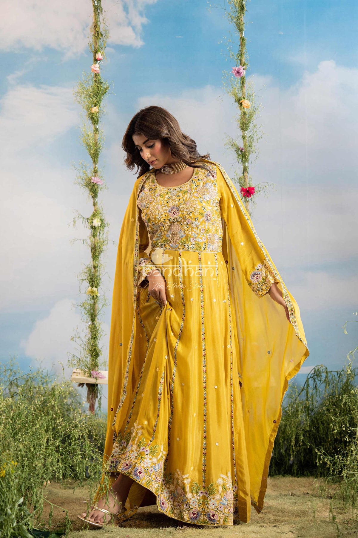 Woman in a yellow traditional outfit with floral patterns standing outdoors on a swing with flowers.