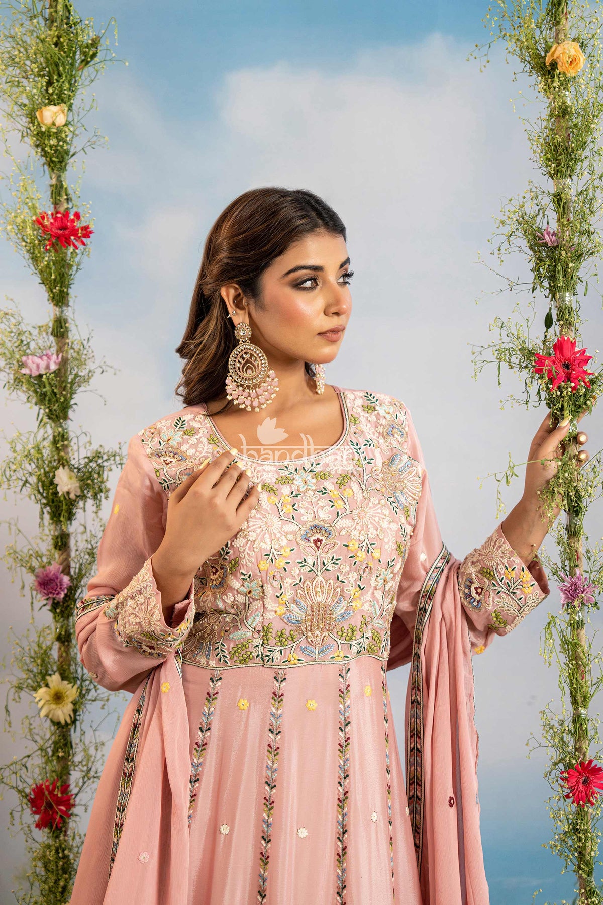 Woman in a pink embroidered traditional outfit standing between floral decorations against a blue sky.