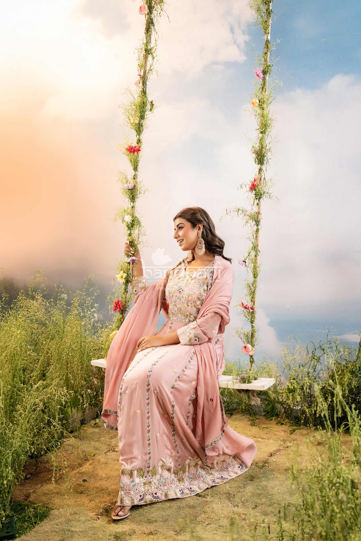 Woman in a pink saree sitting on a swing with floral decorations against a scenic background.