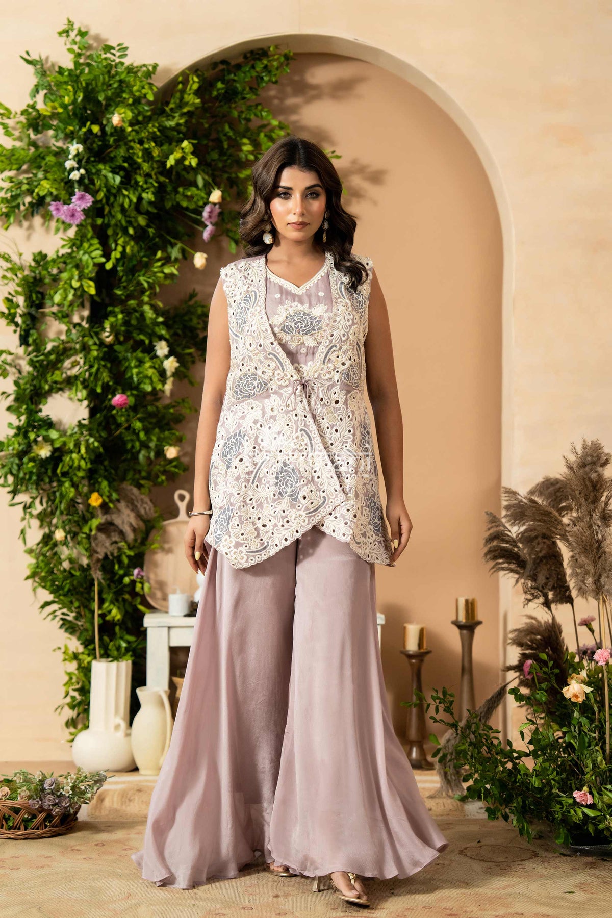 Woman in a sleeveless embroidered top and light purple pants standing in front of an archway with decorative plants.