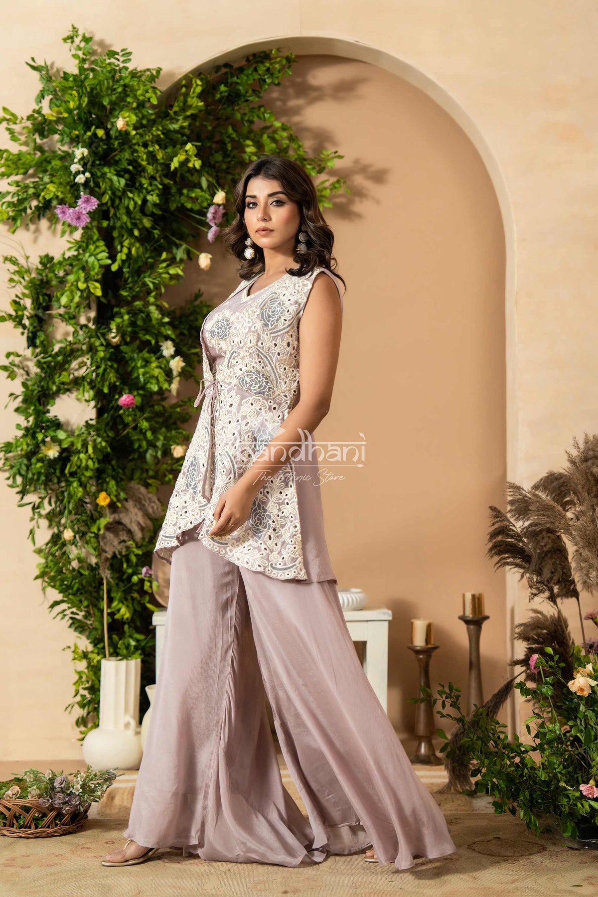 Woman in a floral top and light purple pants standing in front of an arch with greenery.