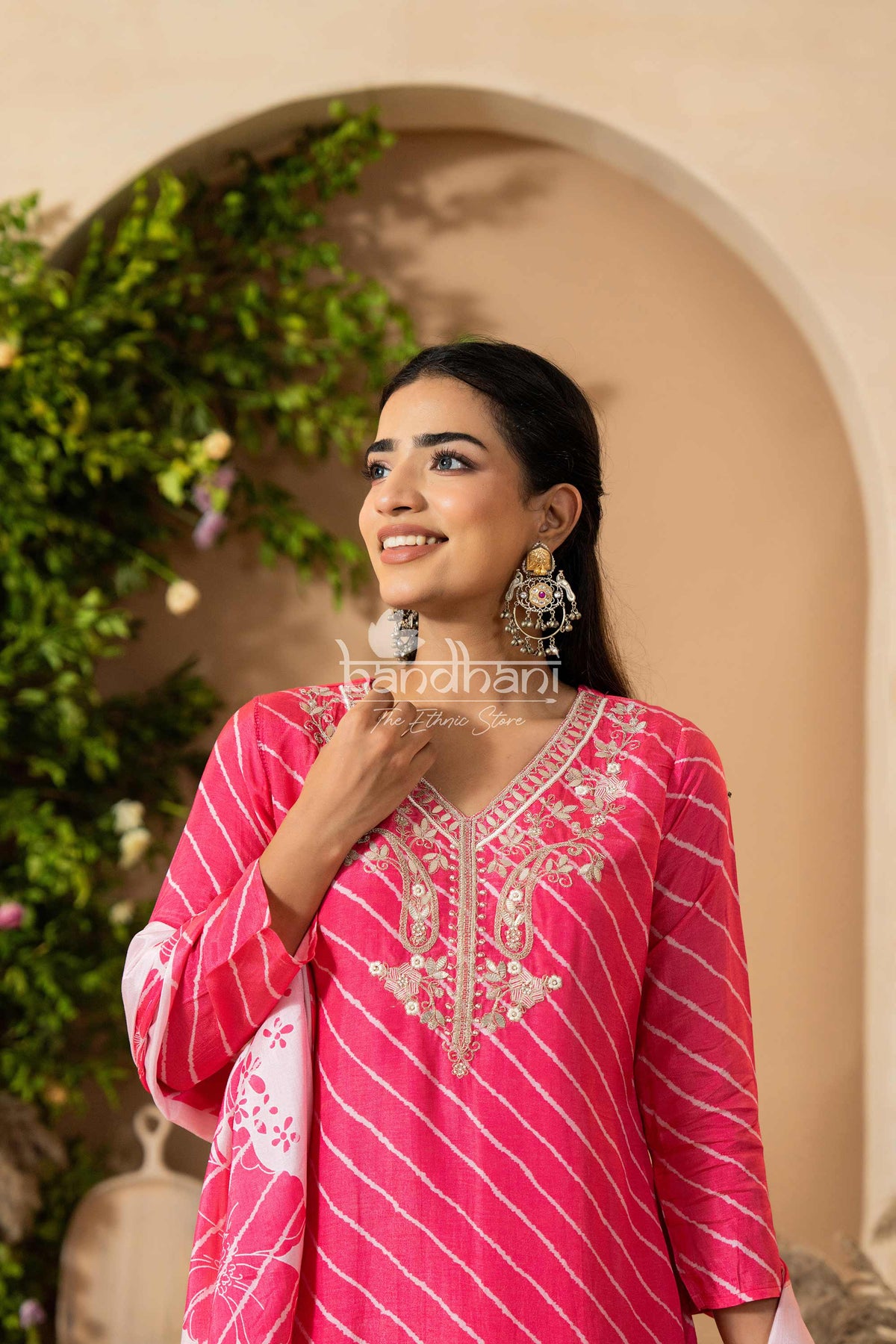 Woman wearing a pink traditional outfit with intricate designs, standing against a decorative background.