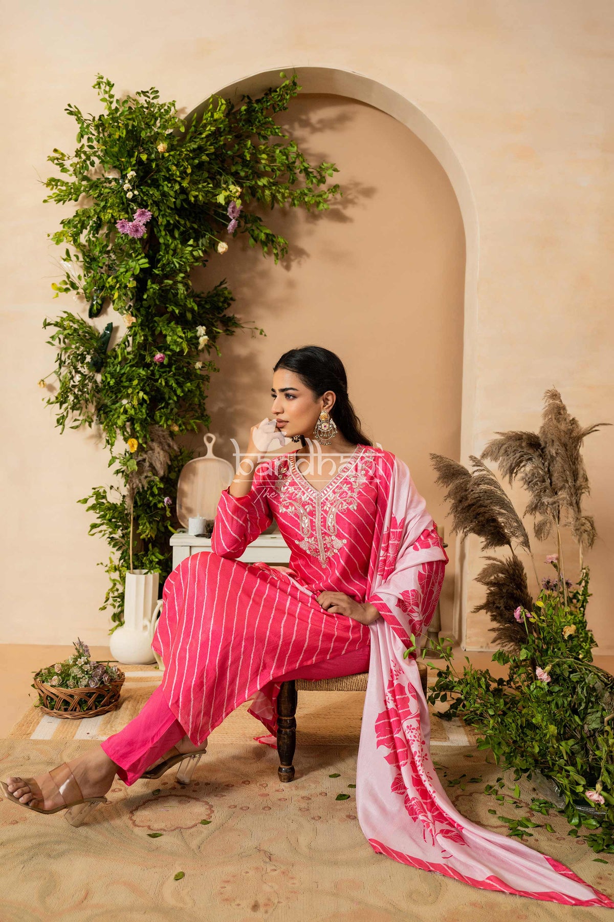 Woman in a pink traditional outfit sitting in a decorated indoor setting with plants and archway.