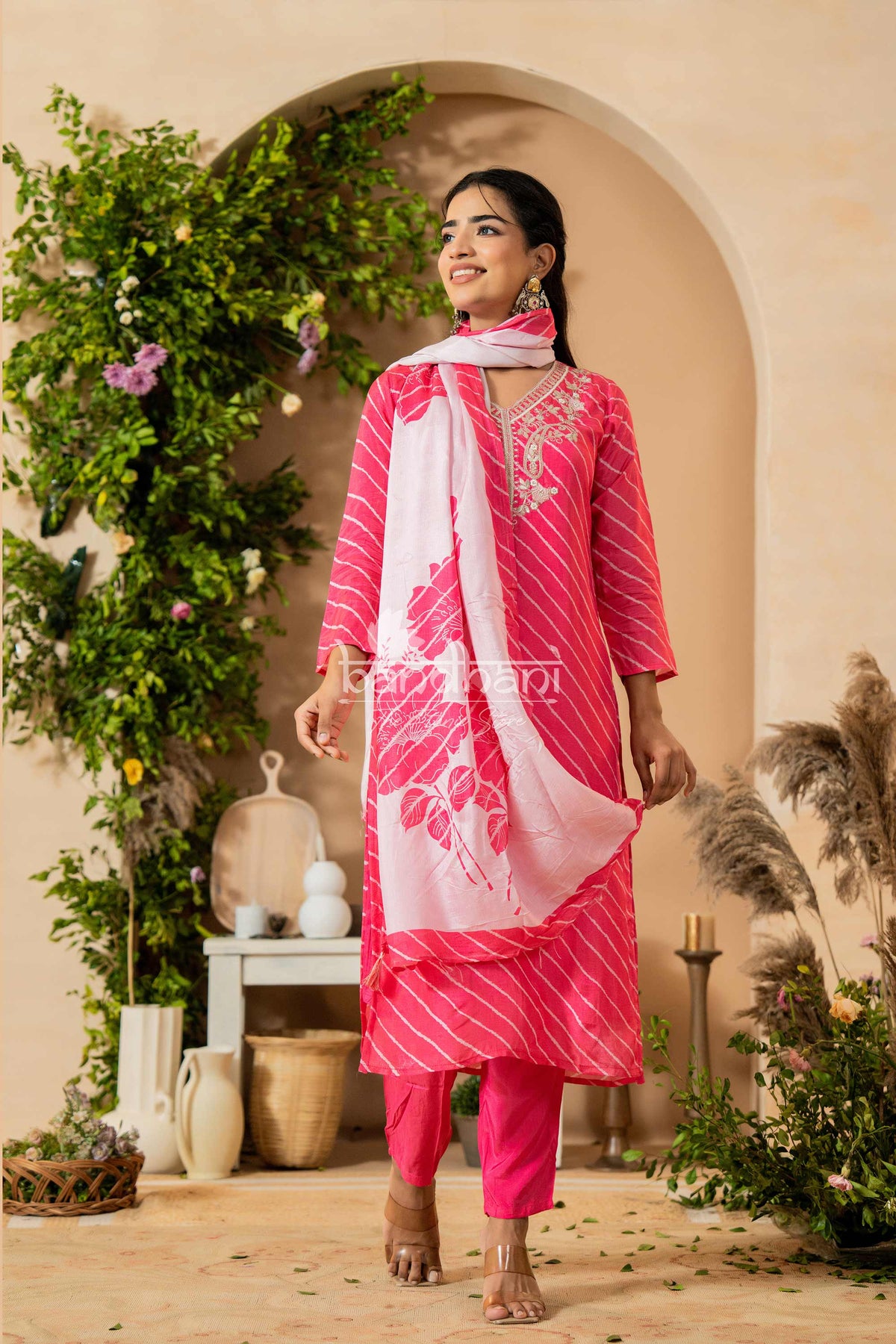 Woman in a pink traditional outfit with a white dupatta standing in a decorated indoor setting.