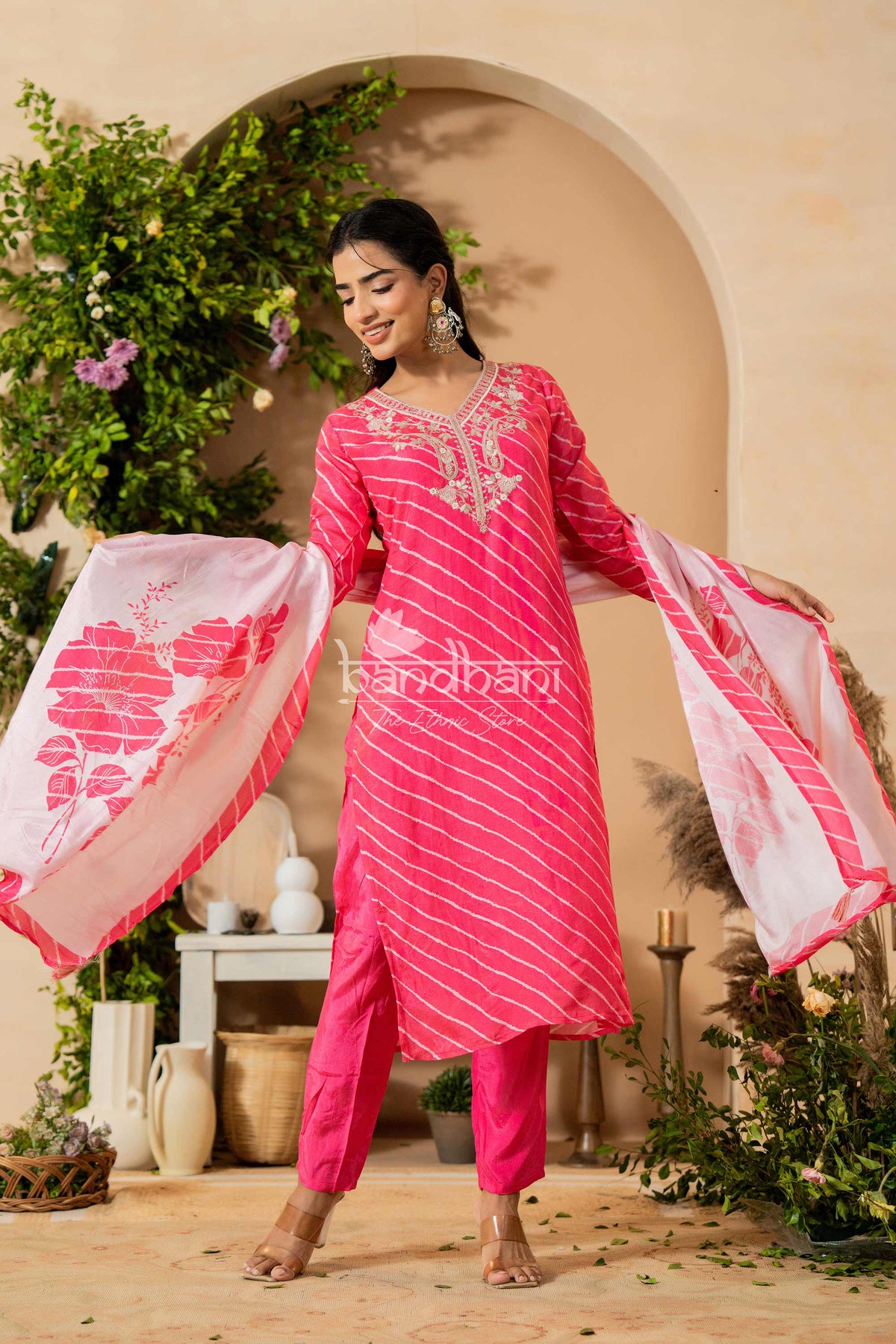 Woman in a pink traditional outfit with a floral design, standing in a decorated indoor setting.