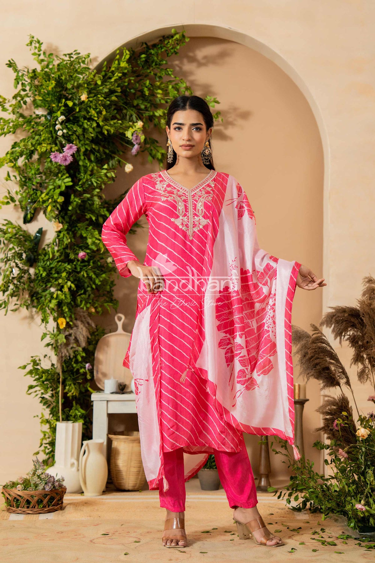 Woman in a pink traditional outfit with a white dupatta standing in front of an archway with greenery.