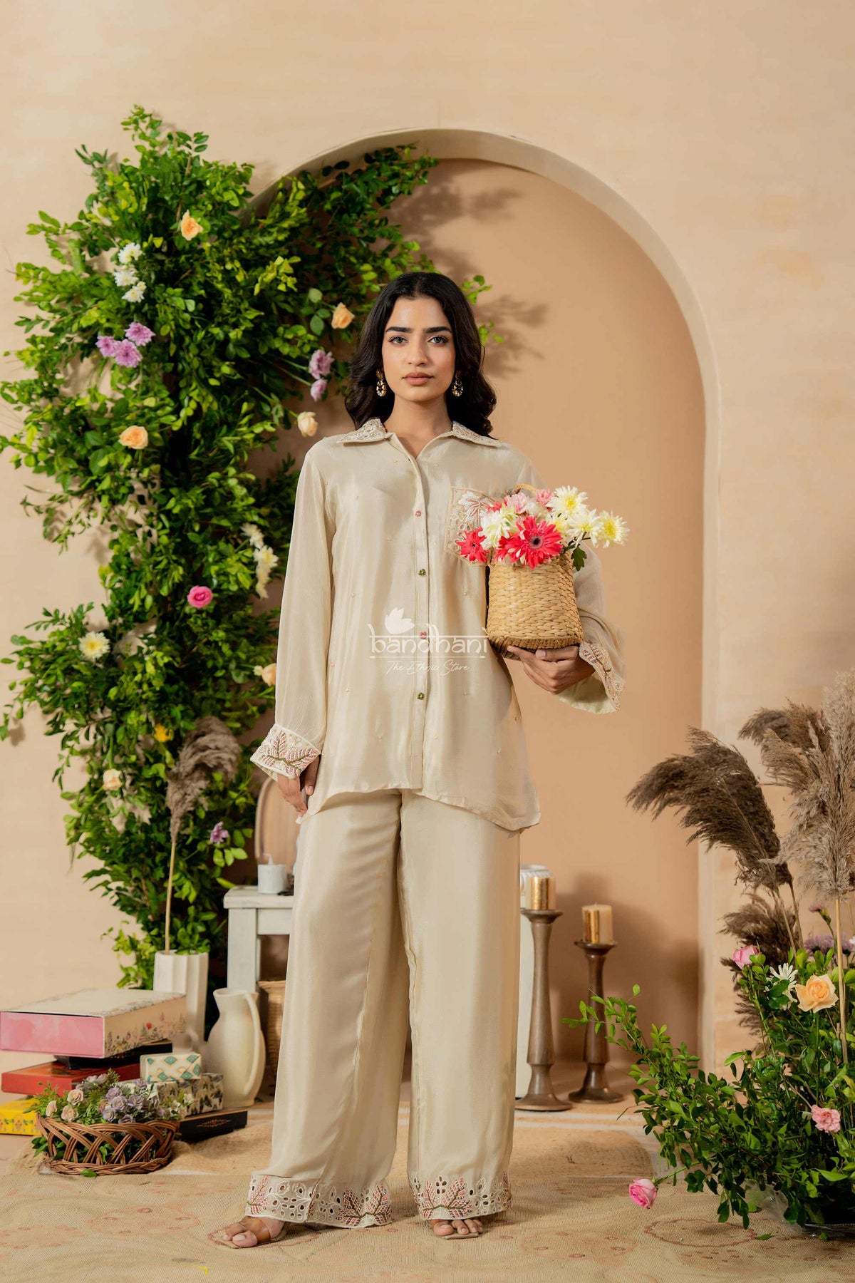 Woman in beige outfit holding flowers in a decorated room with plants and candles.