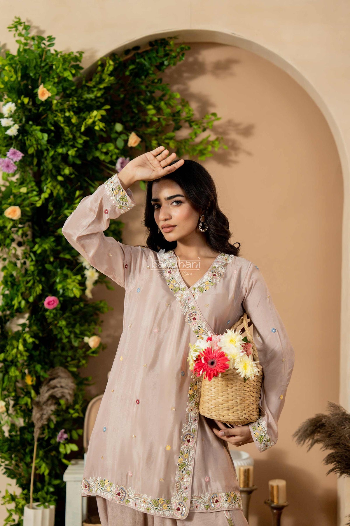 Woman in a light pink traditional outfit holding a basket with flowers, standing against a decorated wall.