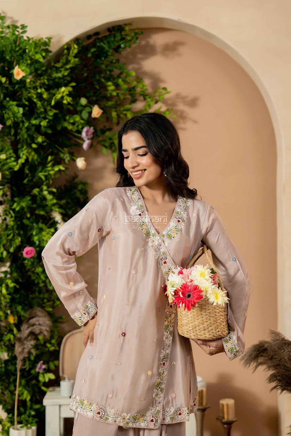 Woman in a light pink traditional outfit holding a basket of flowers, standing against a decorated wall with plants.