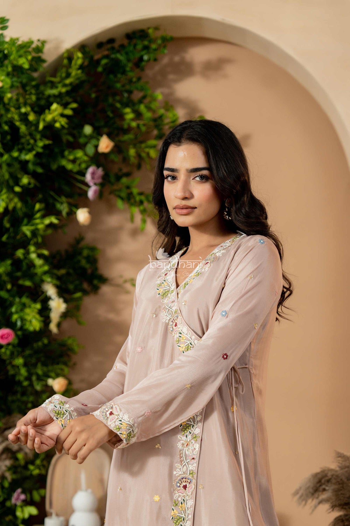 Woman in a light pink embroidered dress standing against a beige wall with greenery.