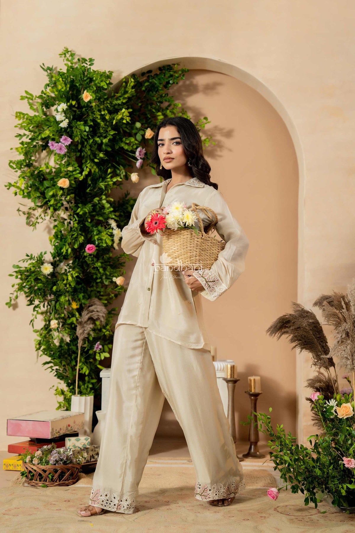 Woman in a beige outfit holding a basket of flowers in a decorated indoor setting with greenery and candles.